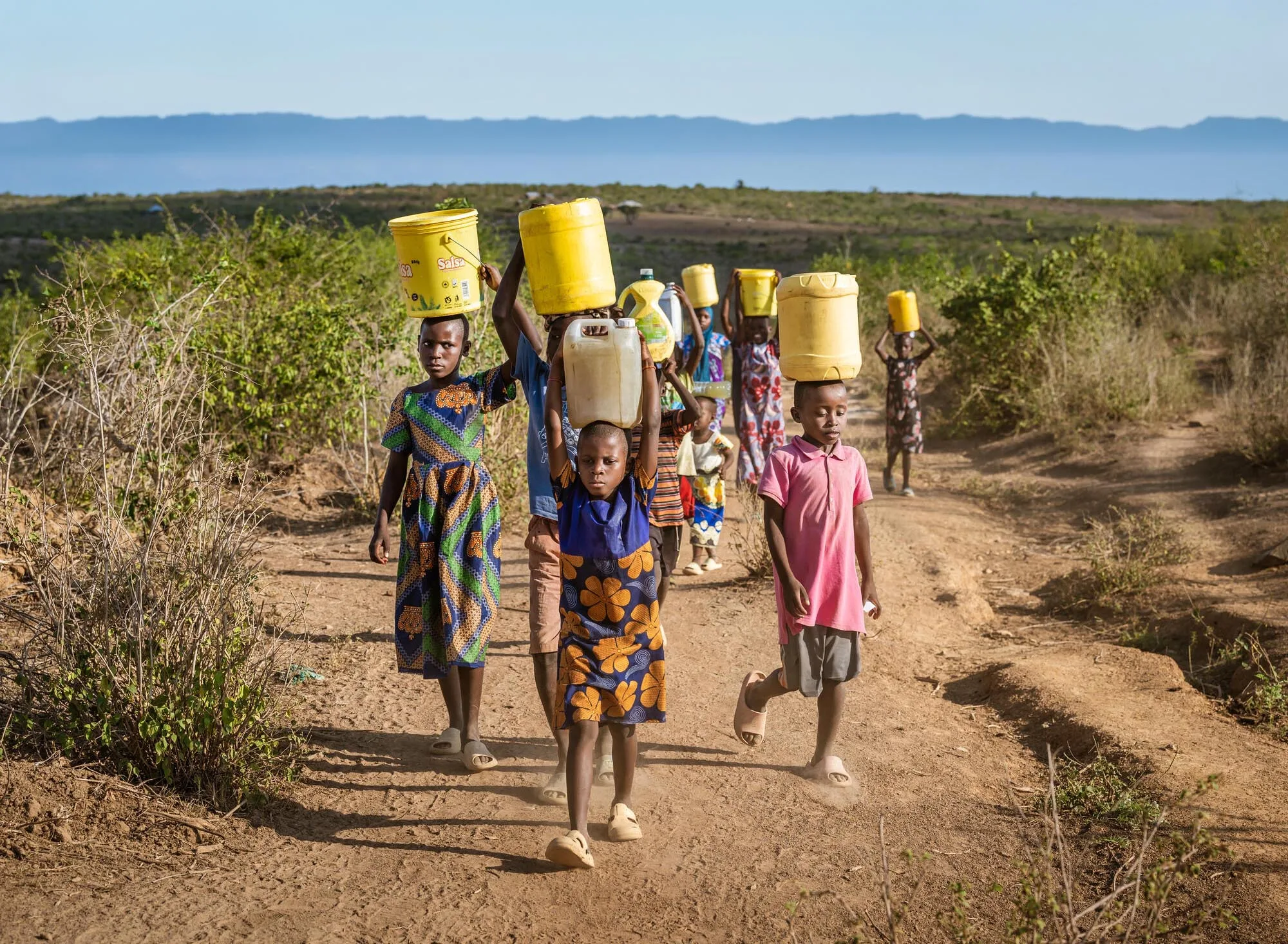 A group of children walk single-file along a dirt path carrying yellow plastic buckets and jerrycans on their heads, with dry brush and distant hills in the bac