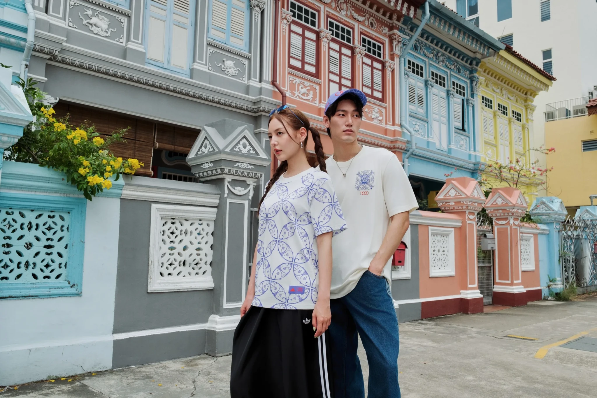 A young woman and man in patterned adidas T-shirts and denim pose in front of colourful shophouses with ornate facades
