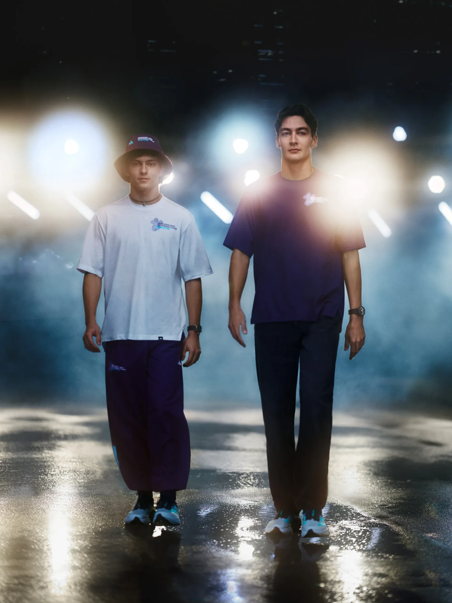Two young men in Mercedes-AMG PETRONAS F1 Team streetwear, one in a purple bucket hat, stand on a wet, moodily lit surface