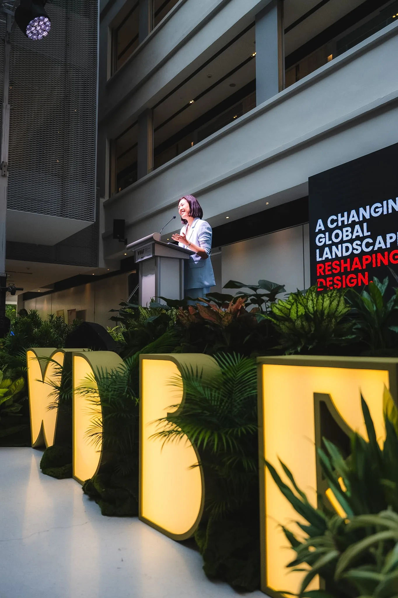 A speaker at a podium in front of a screen reading Reshaping Design