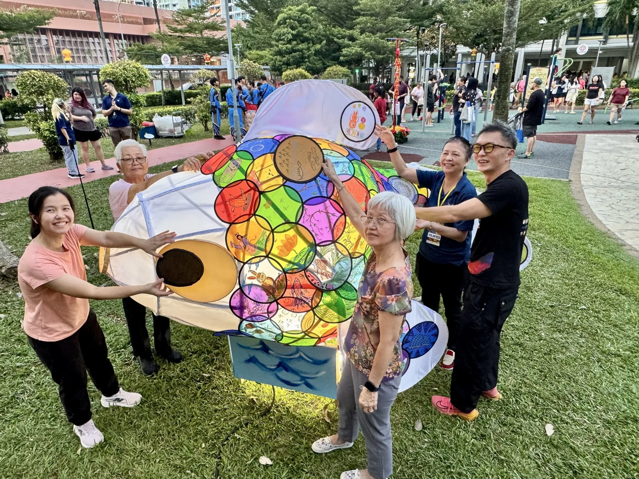 Visitors posing with a large colourful stained-glass-style community art installation in an urban plaza
