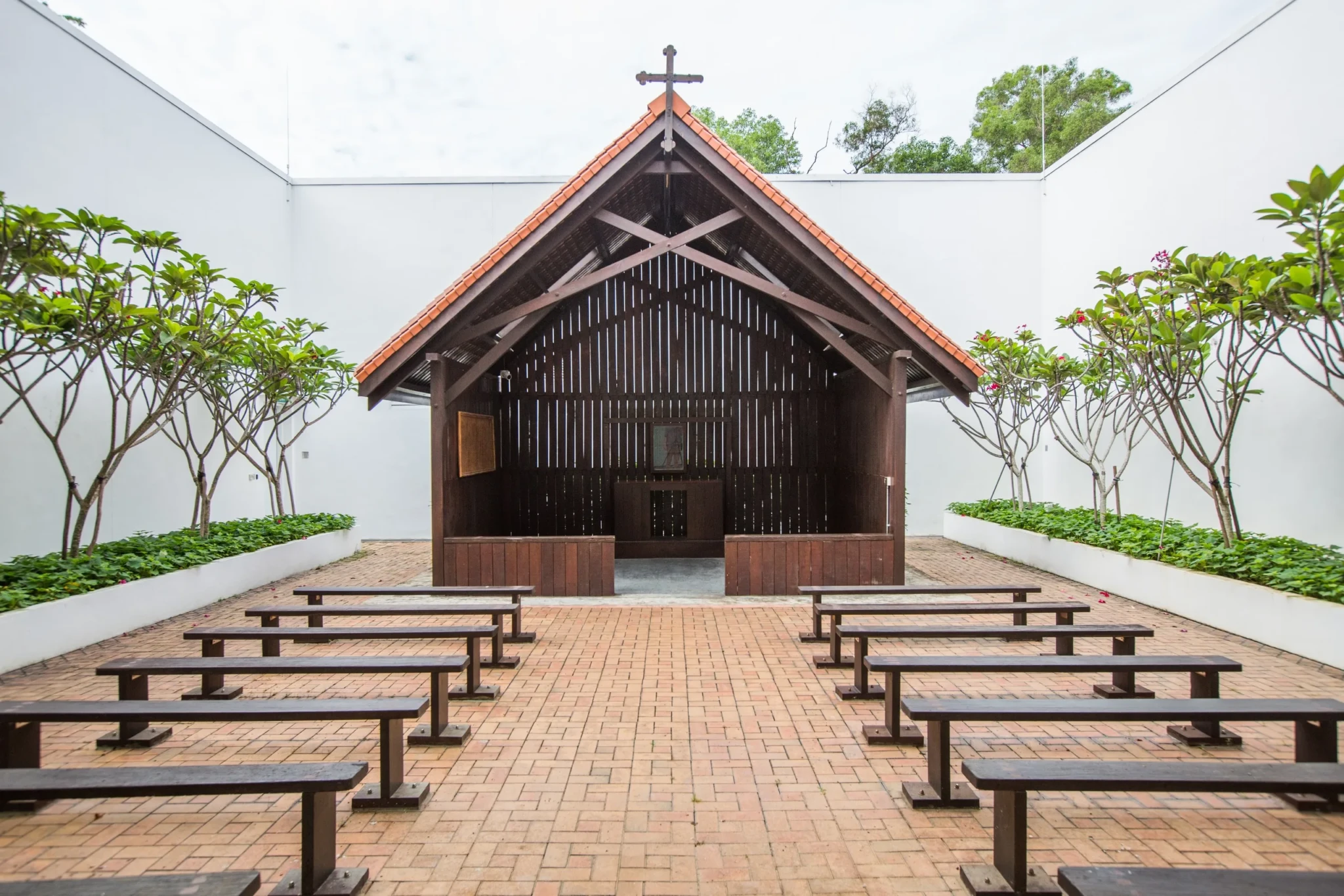 Modern chapel with wooden slat walls, orange roof and cross, wooden benches in courtyard with landscaping