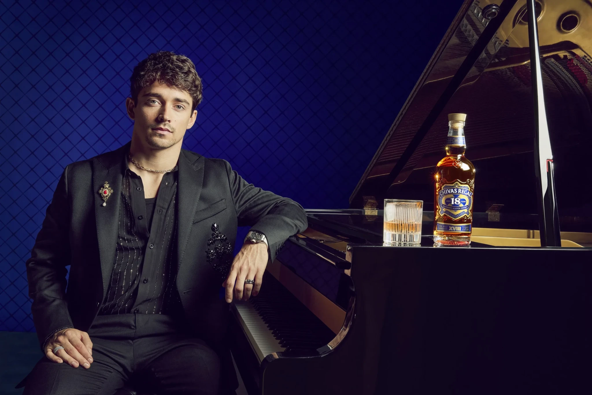 Charles Leclerc sits beside a grand piano with a glass of whisky and a bottle of Chivas Regal 18 on it, against a blue backdrop
