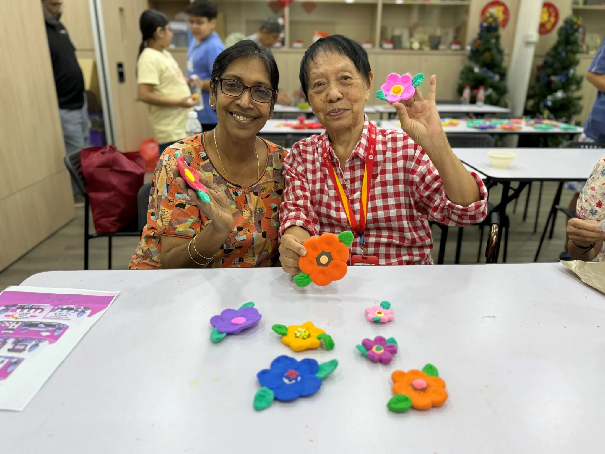 Two smiling workshop participants holding colorful air-dry clay flowers they created at a craft booth.