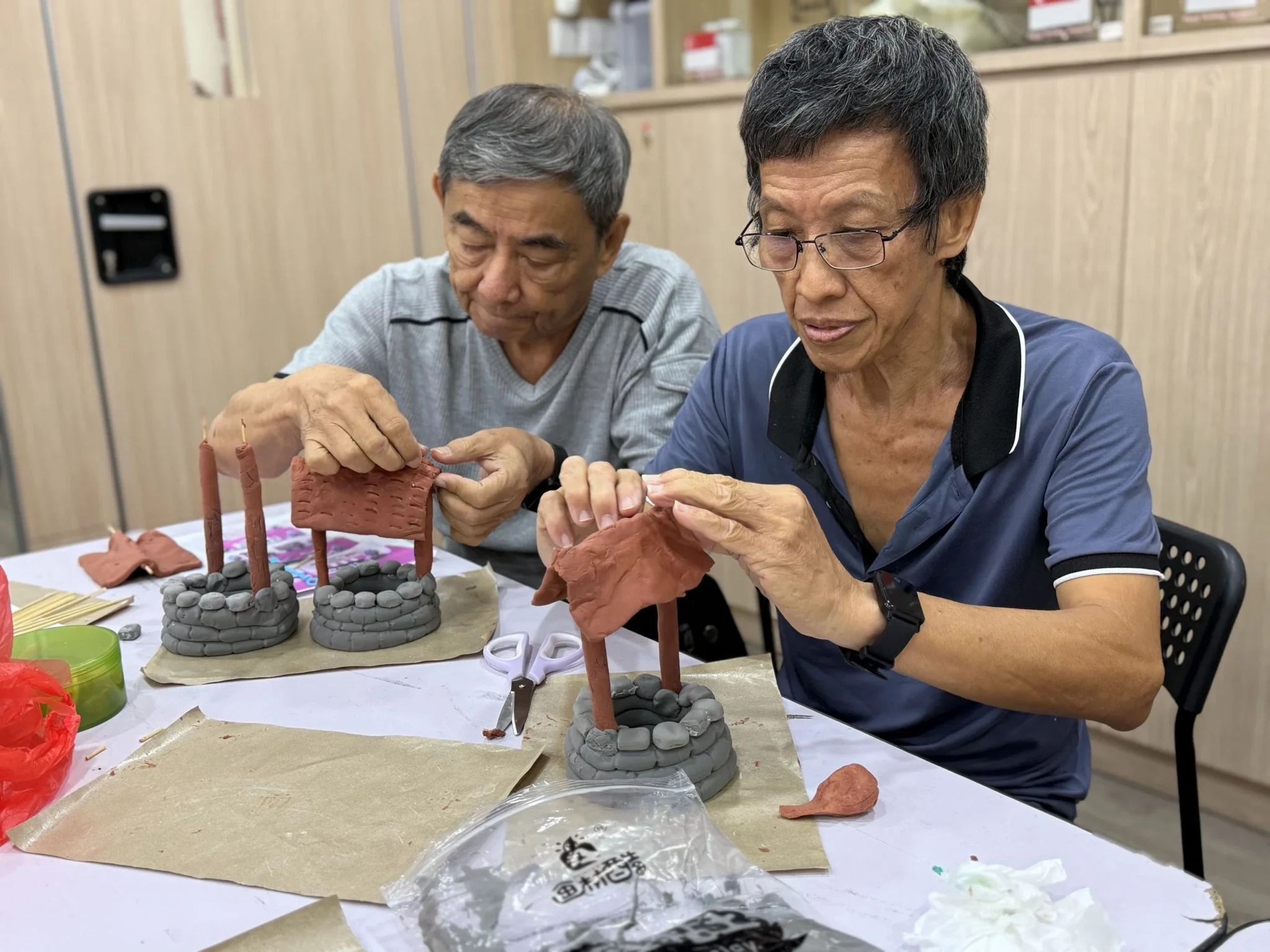 Two adults working together on small clay architectural structures at a craft workshop table