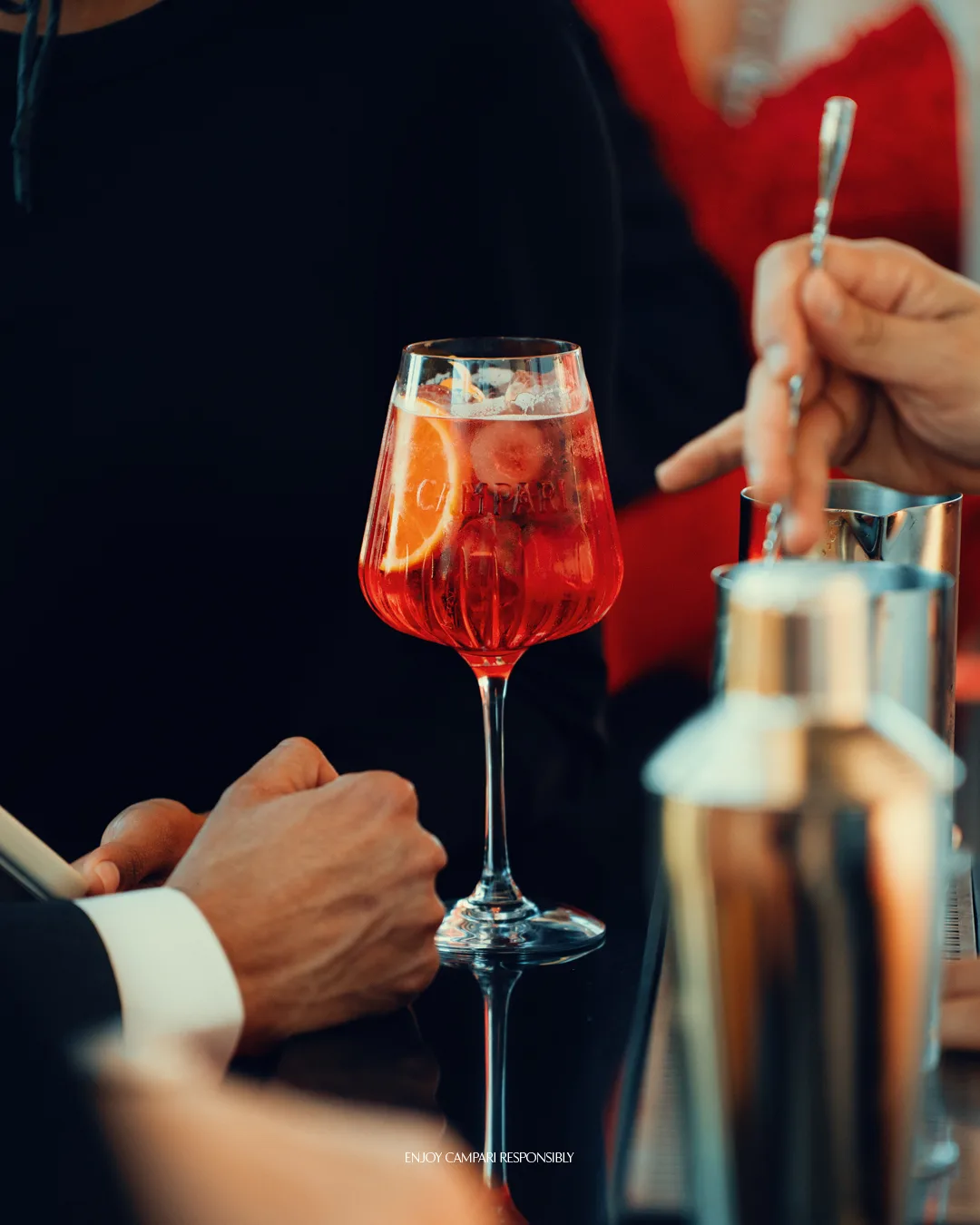 A person holding a Campari Spritz with ice and an orange slice at a social gathering