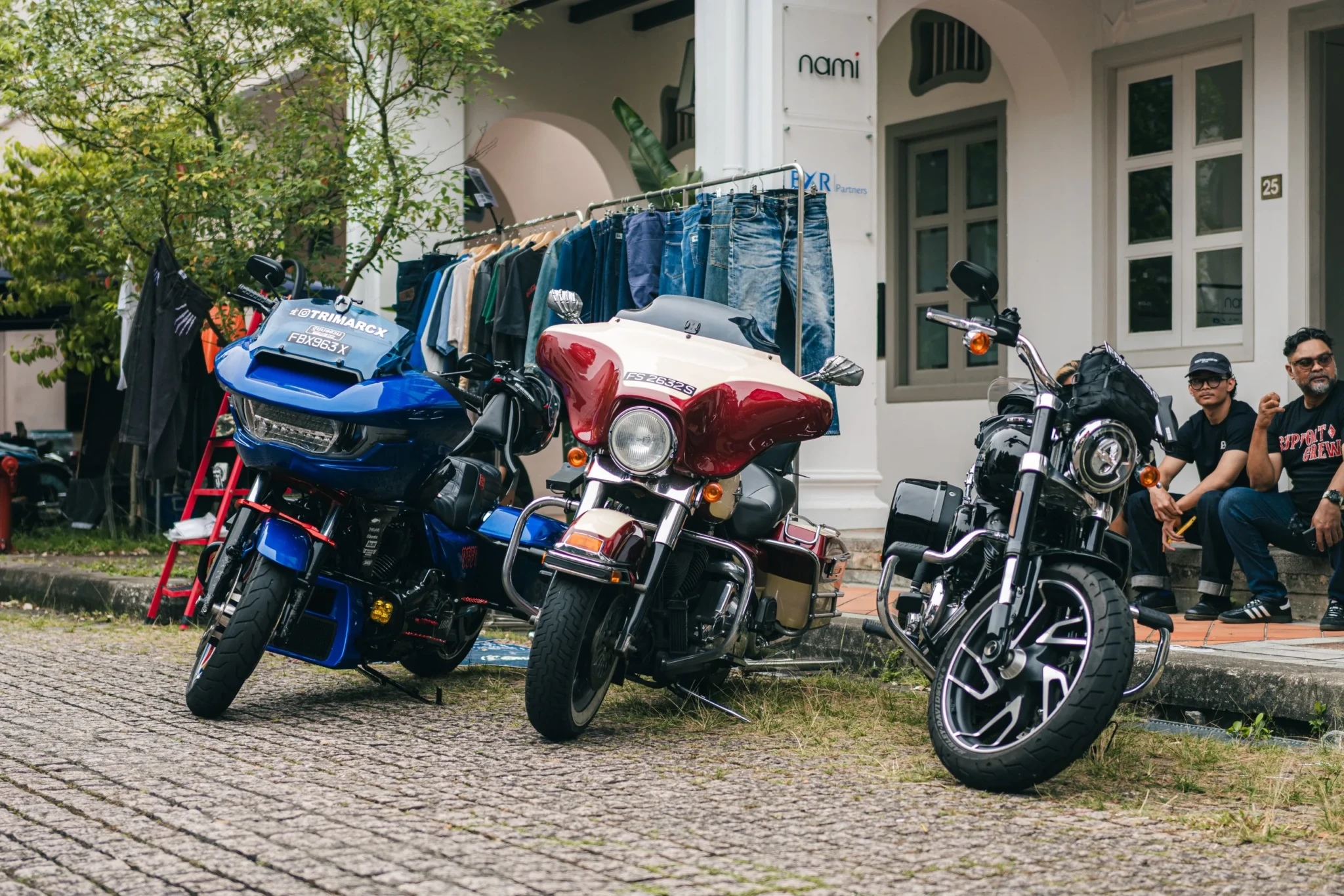 Three motorcycles parked in a row on a cobbled street, with a rack of clothes and two people sitting on a step behind them