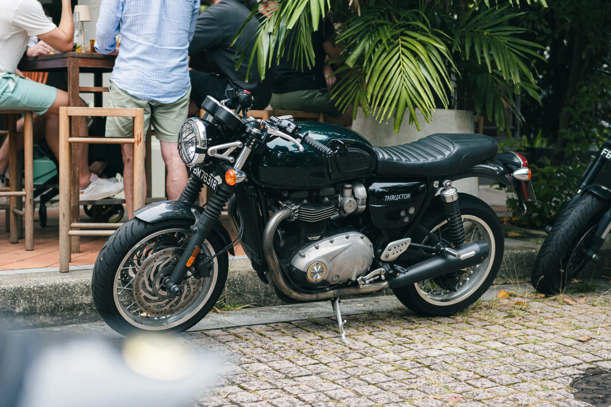 Custom black Triumph cafe racer motorcycle on display with tropical plants and spectators in background