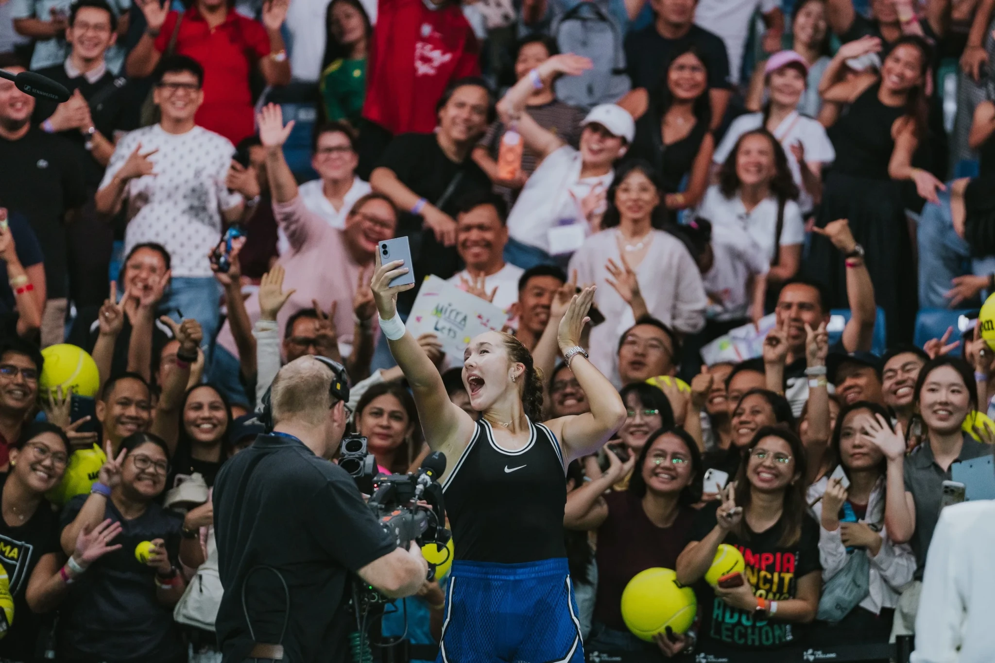 Tennis player Mirra Andreeva holding up a phone amid a cheering crowd at an indoor venue