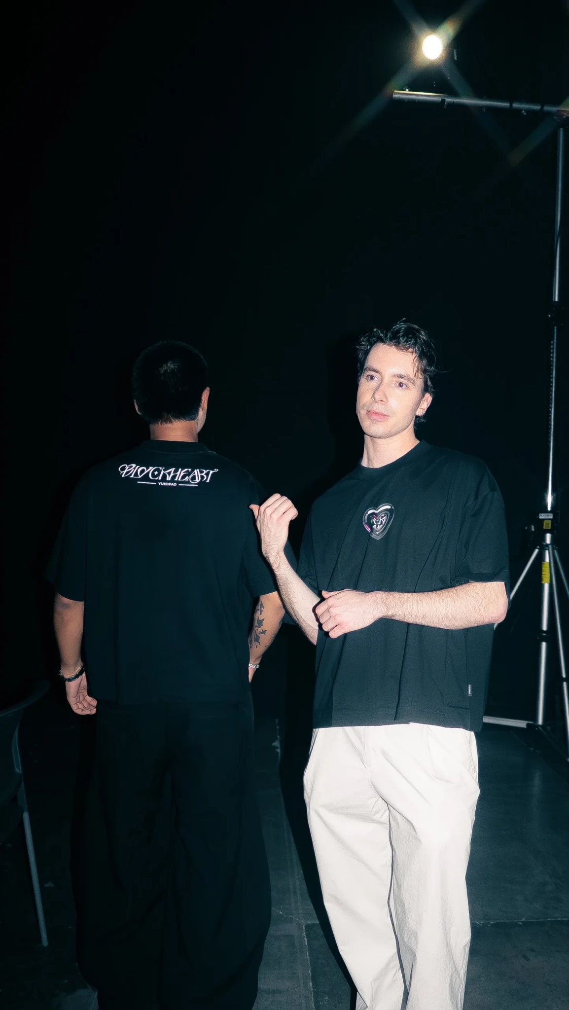 Two men in black t-shirts against a dark background, one facing away from the camera