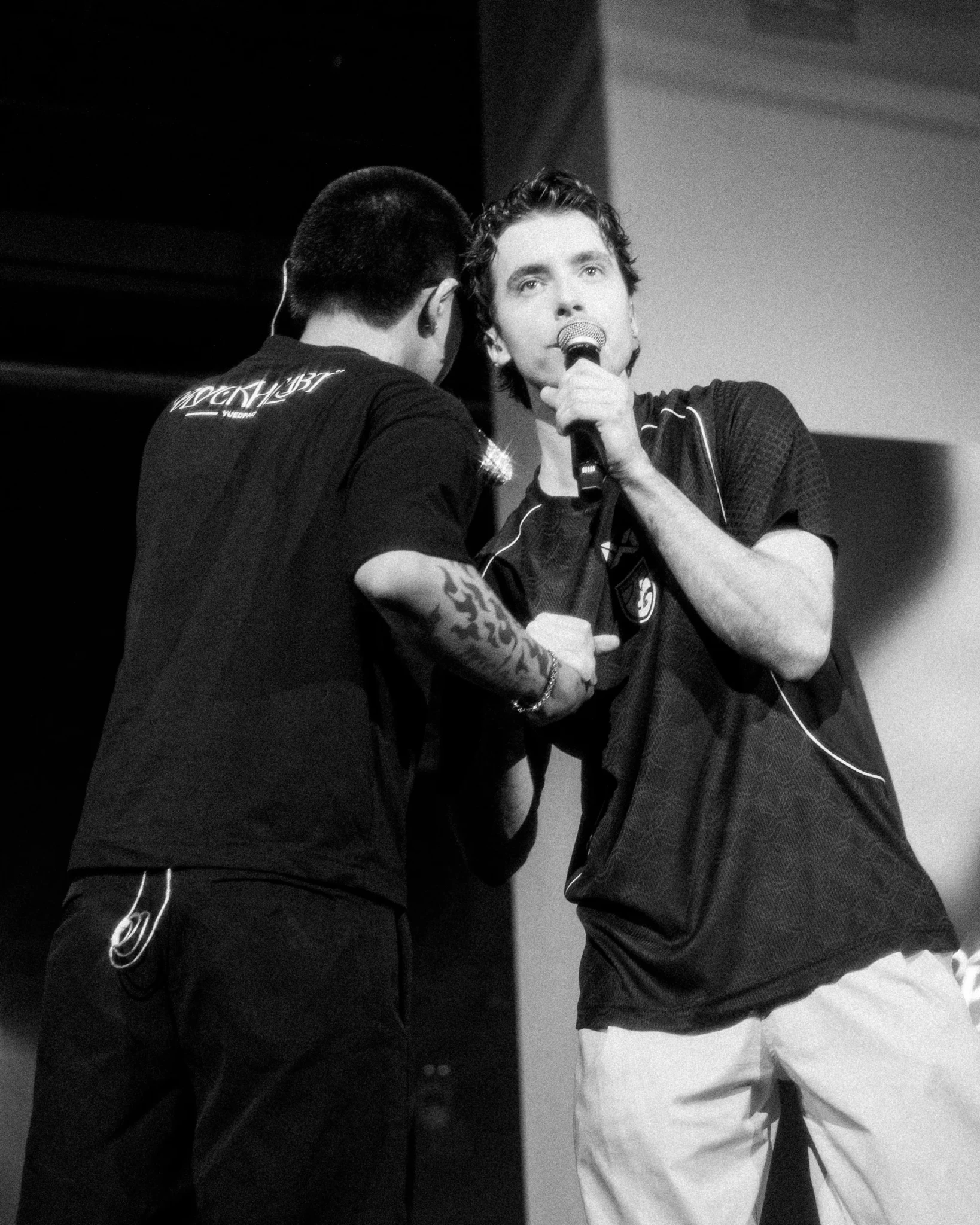 Two men in a black-and-white photograph, one holding a microphone and the other turned away