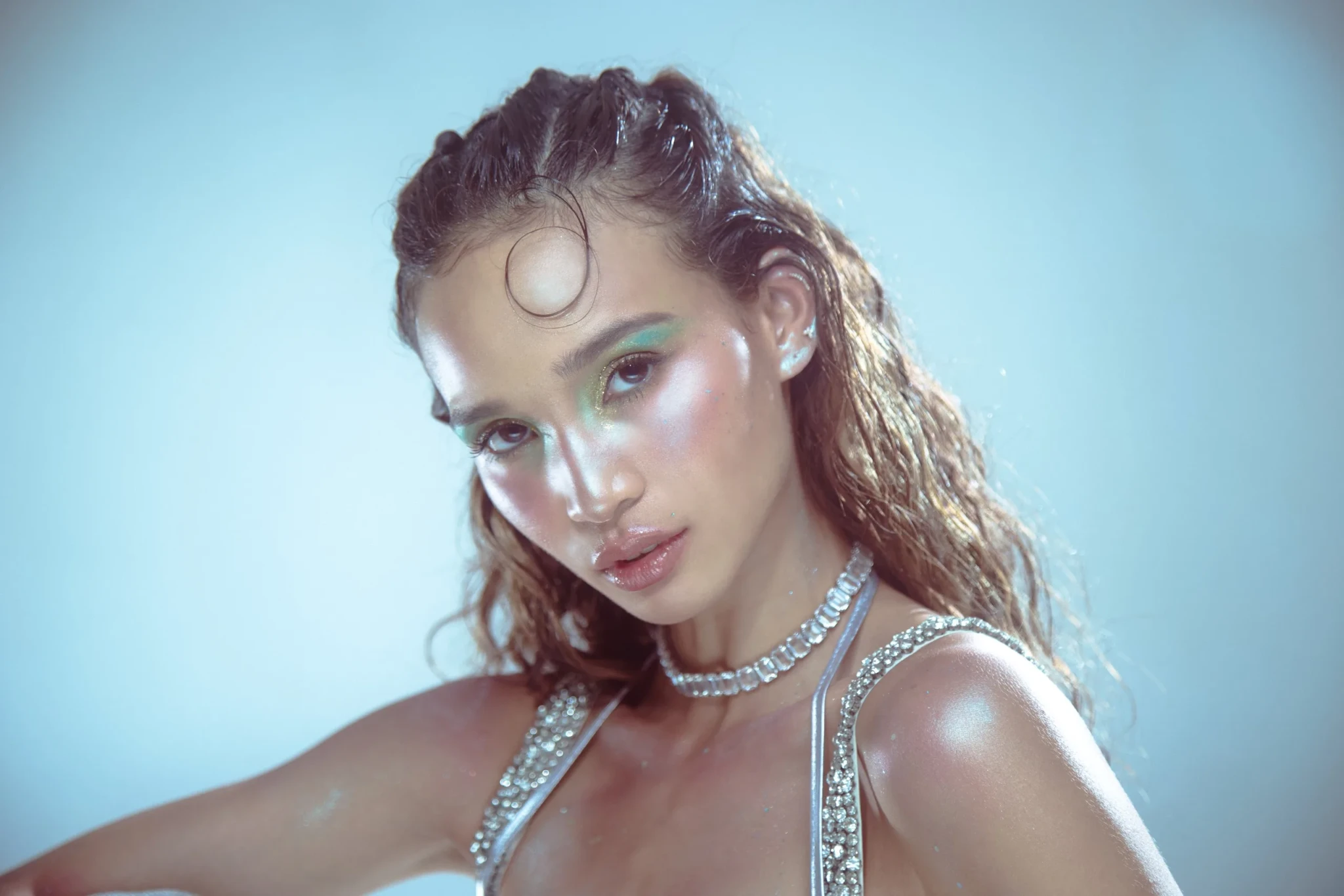 Iman Fandi in a portrait with green eyeshadow and a crystal-embellished top against a pale blue background