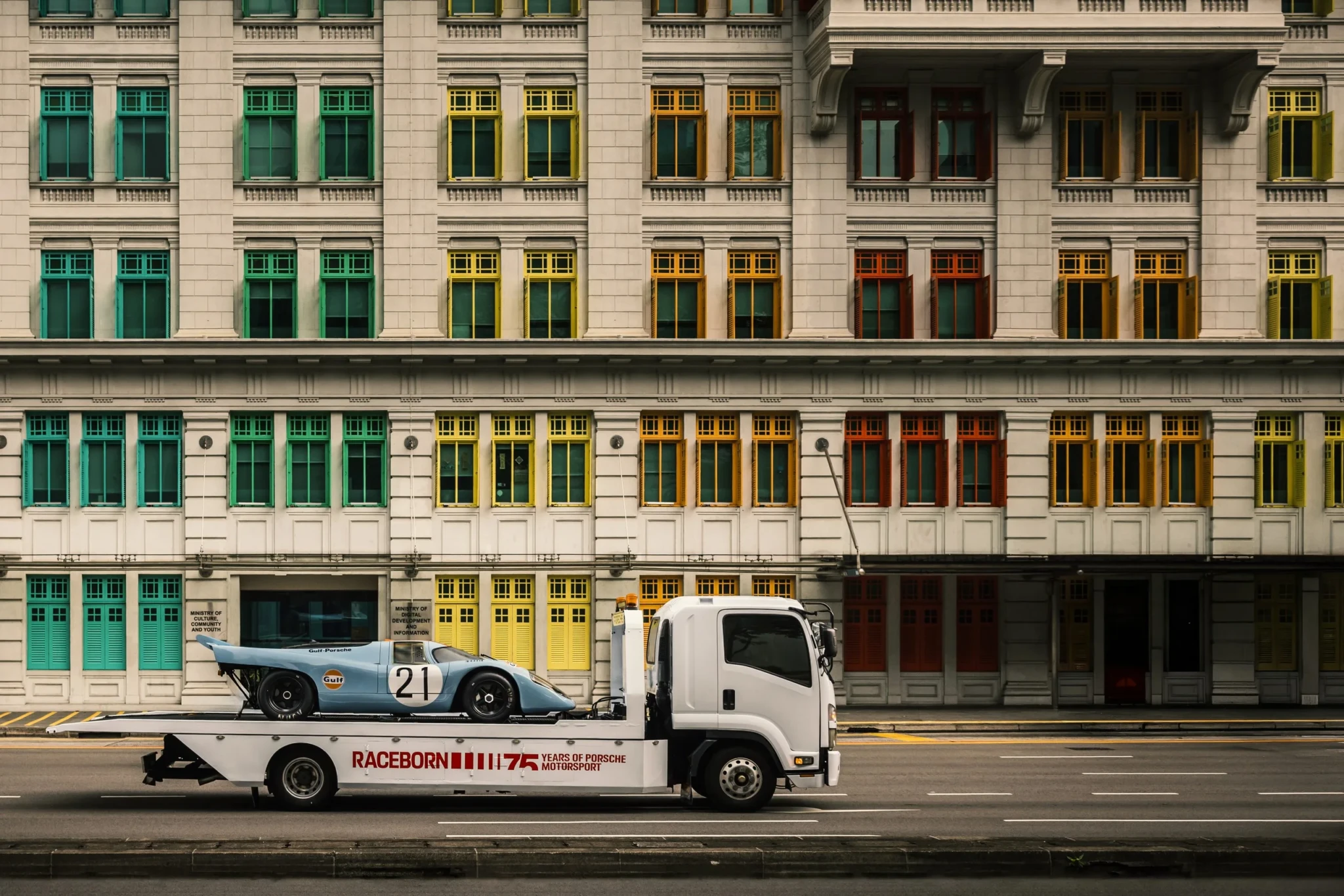 A Raceborn-branded flatbed truck carrying the Porsche 917 KH past a historic Singapore building