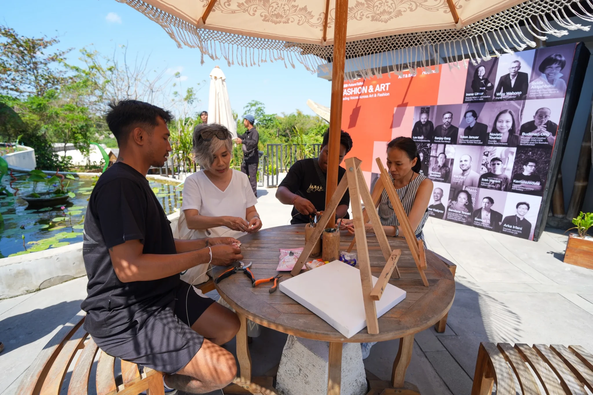 Four people working with wooden crafting tools at a table under a shaded structure during the Art & Bali 2026 event.