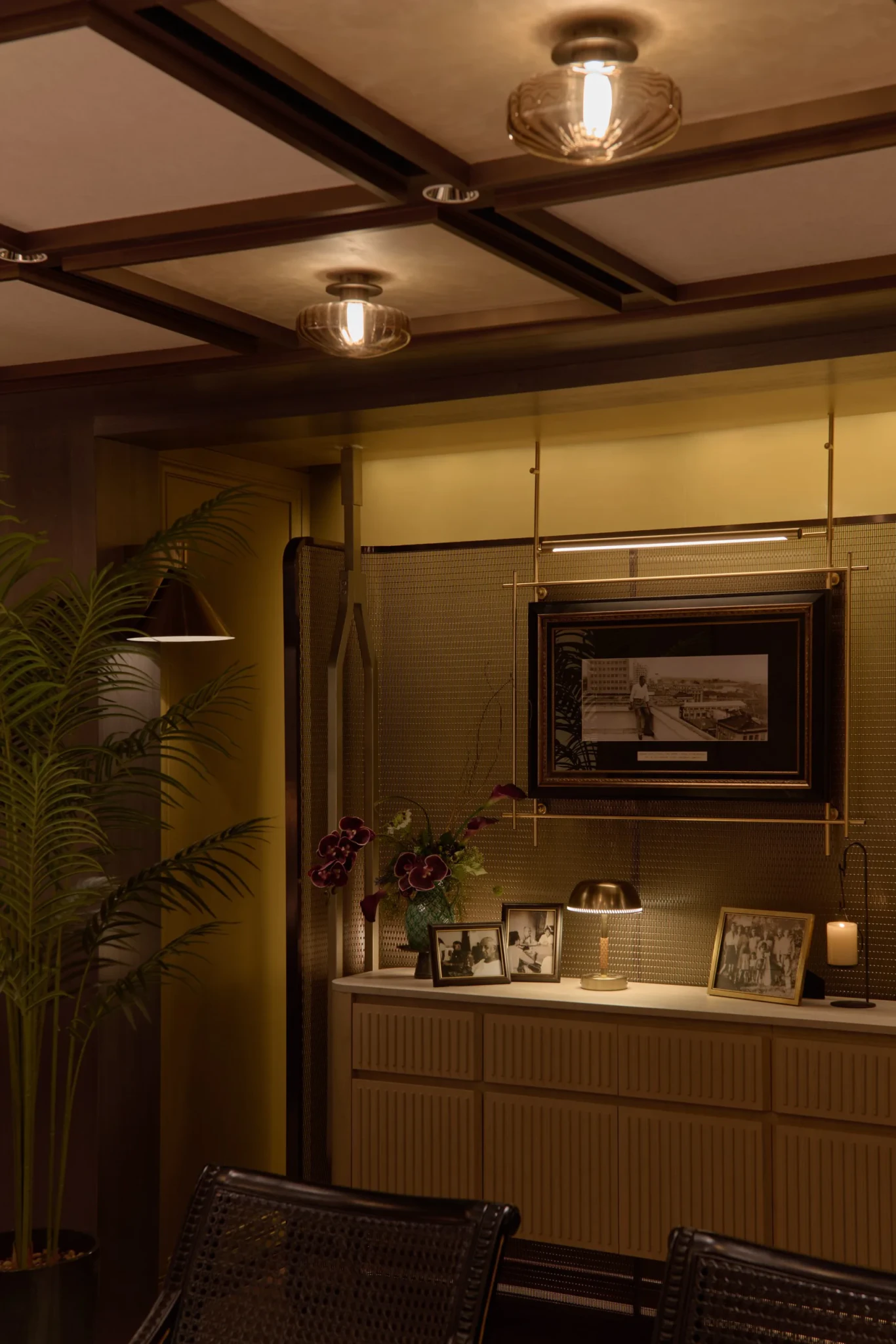 Restaurant dining room with dark wooden ceiling beams, cream walls, lit sideboard with photo displays, framed art, and floral arrangement.