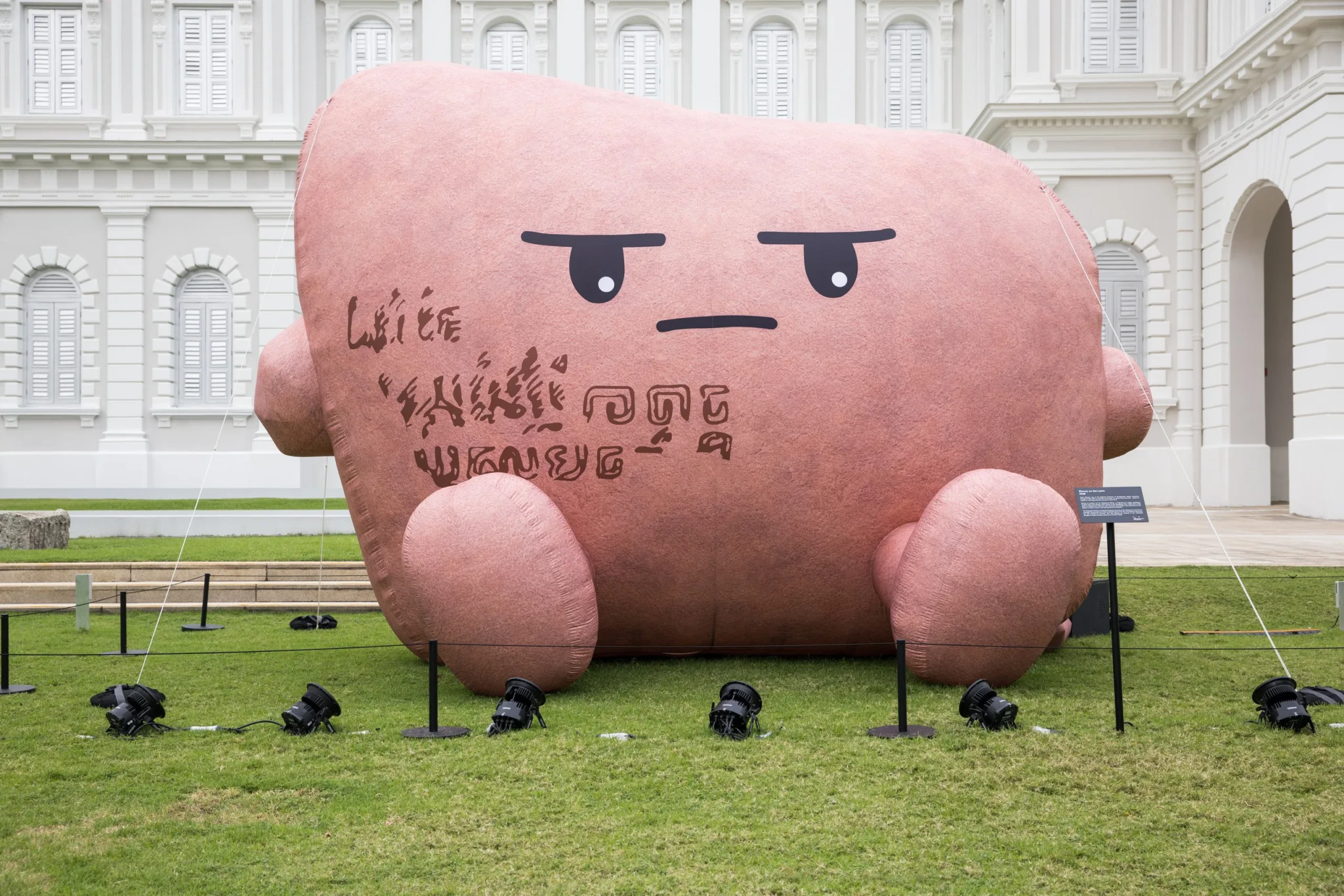 Large pink cartoon-like sculpture with downturned eyes on museum lawn