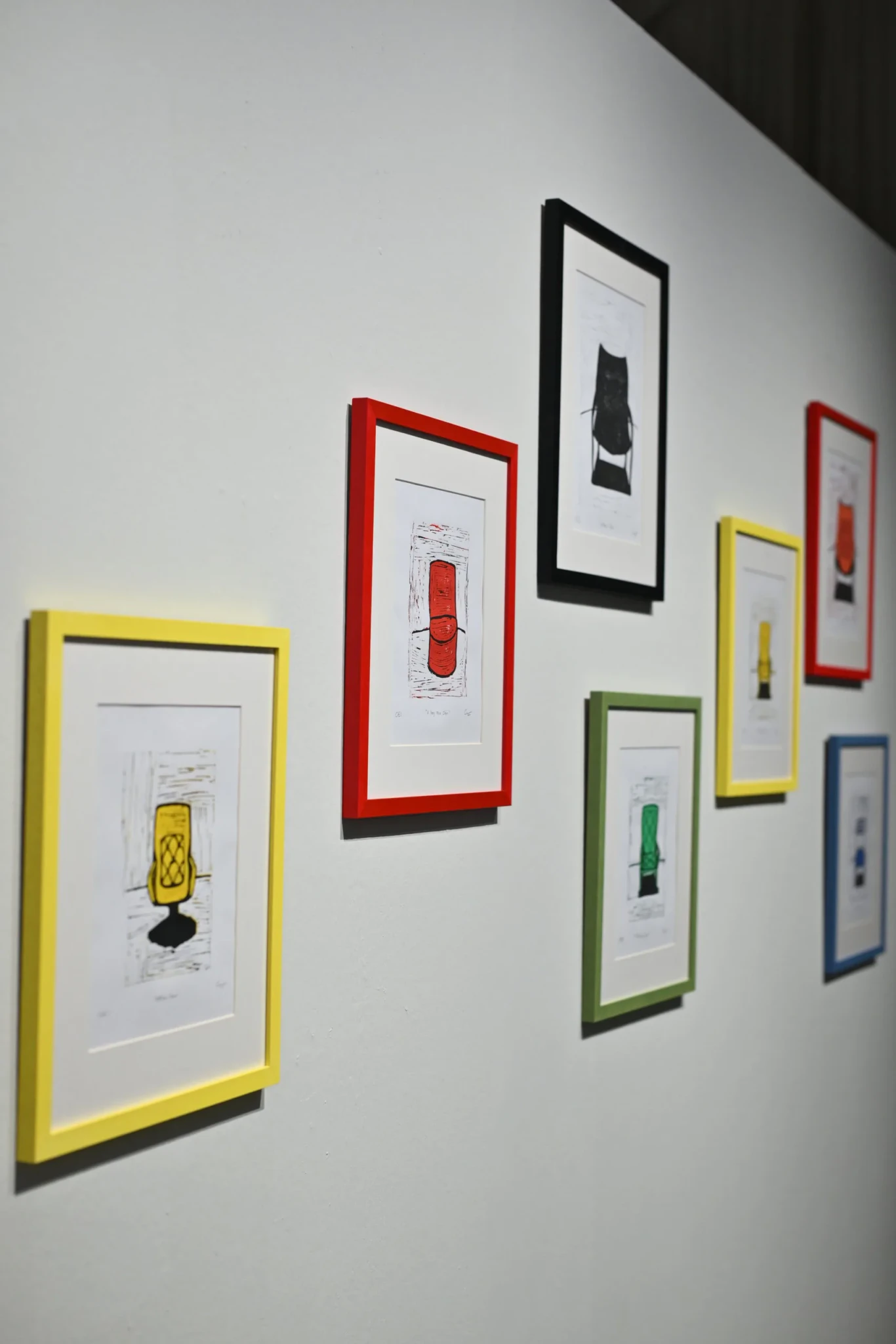 Framed linocut prints of everyday objects in yellow, red, black, green and blue frames on a gallery wall