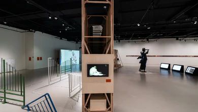 Wide view of the SAMCollection: The Present is Unstable gallery with a tall wooden structure, video displays and metal barriers