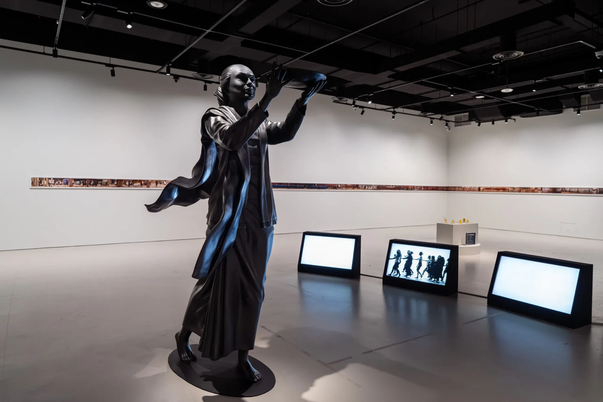 A dark sculptural figure with raised arms beside screens in Nadiah Bamadhaj's Casting Spells for the Movement