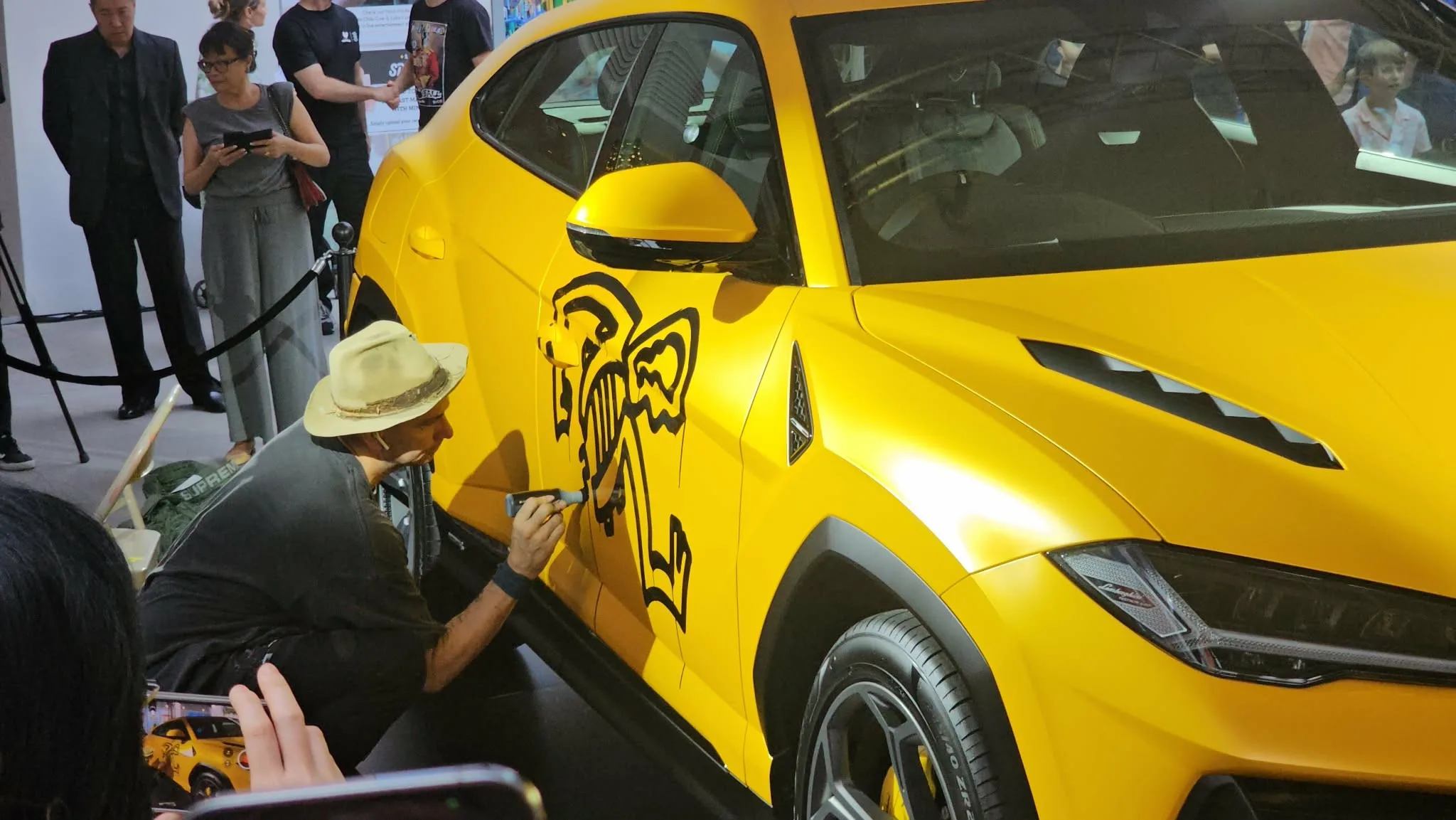 Danny Minnick painting his figures onto a yellow Lamborghini Urus S as a crowd watches