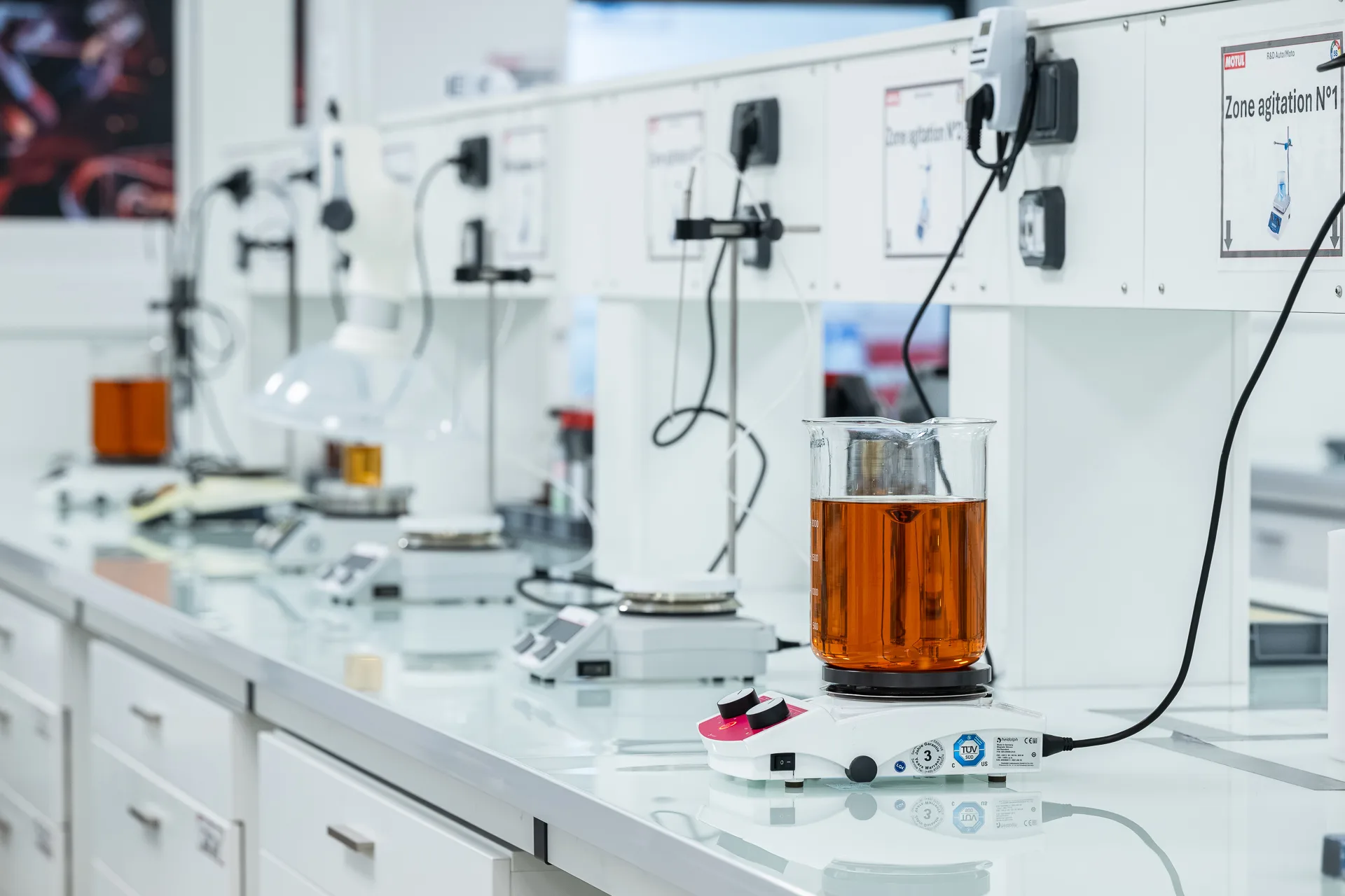 Orange gear oil in a beaker on a magnetic stirrer in a laboratory testing setup with white equipment and cables in the background