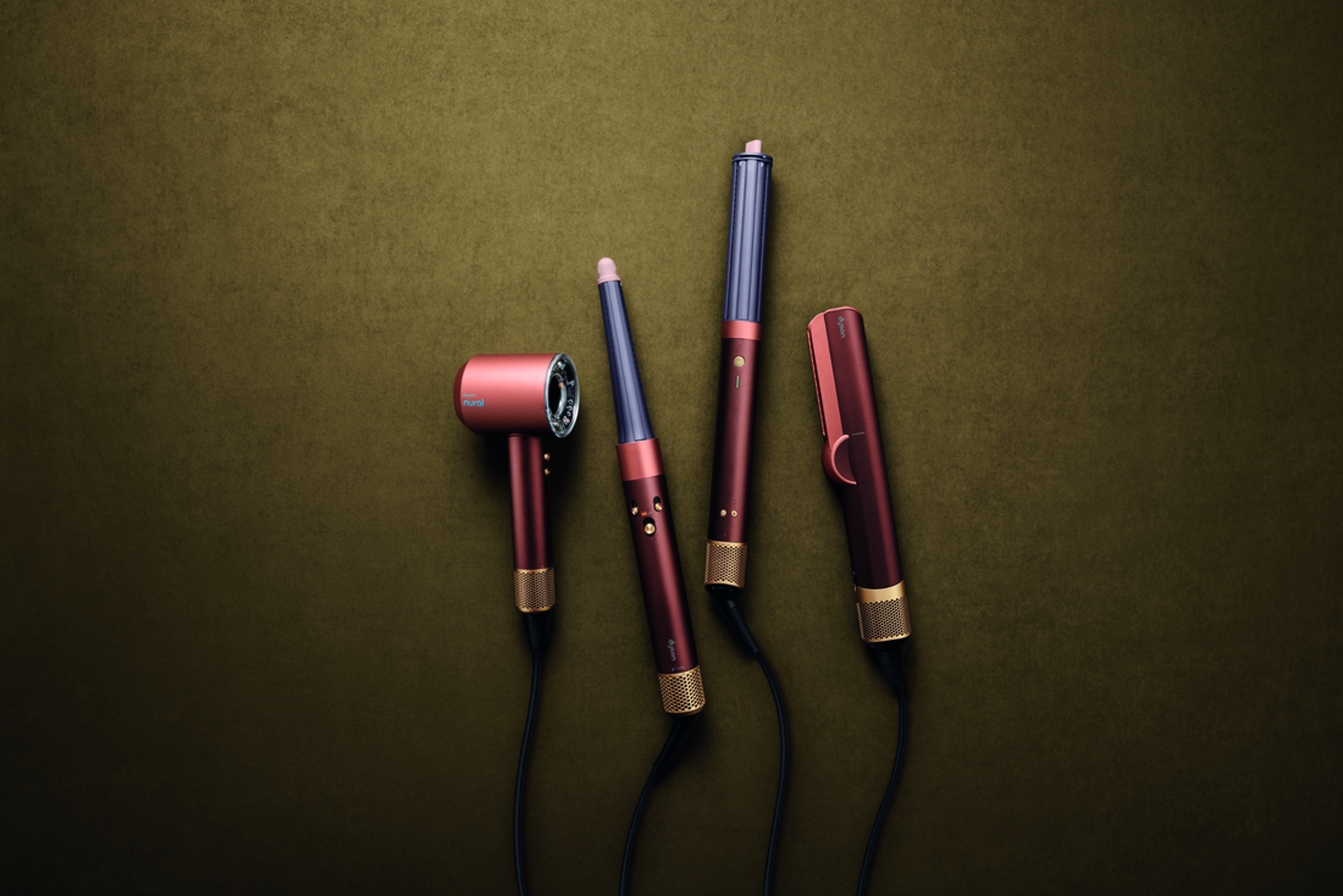 Five Dyson hair care devices in plum, copper, and gold tones arranged on olive background