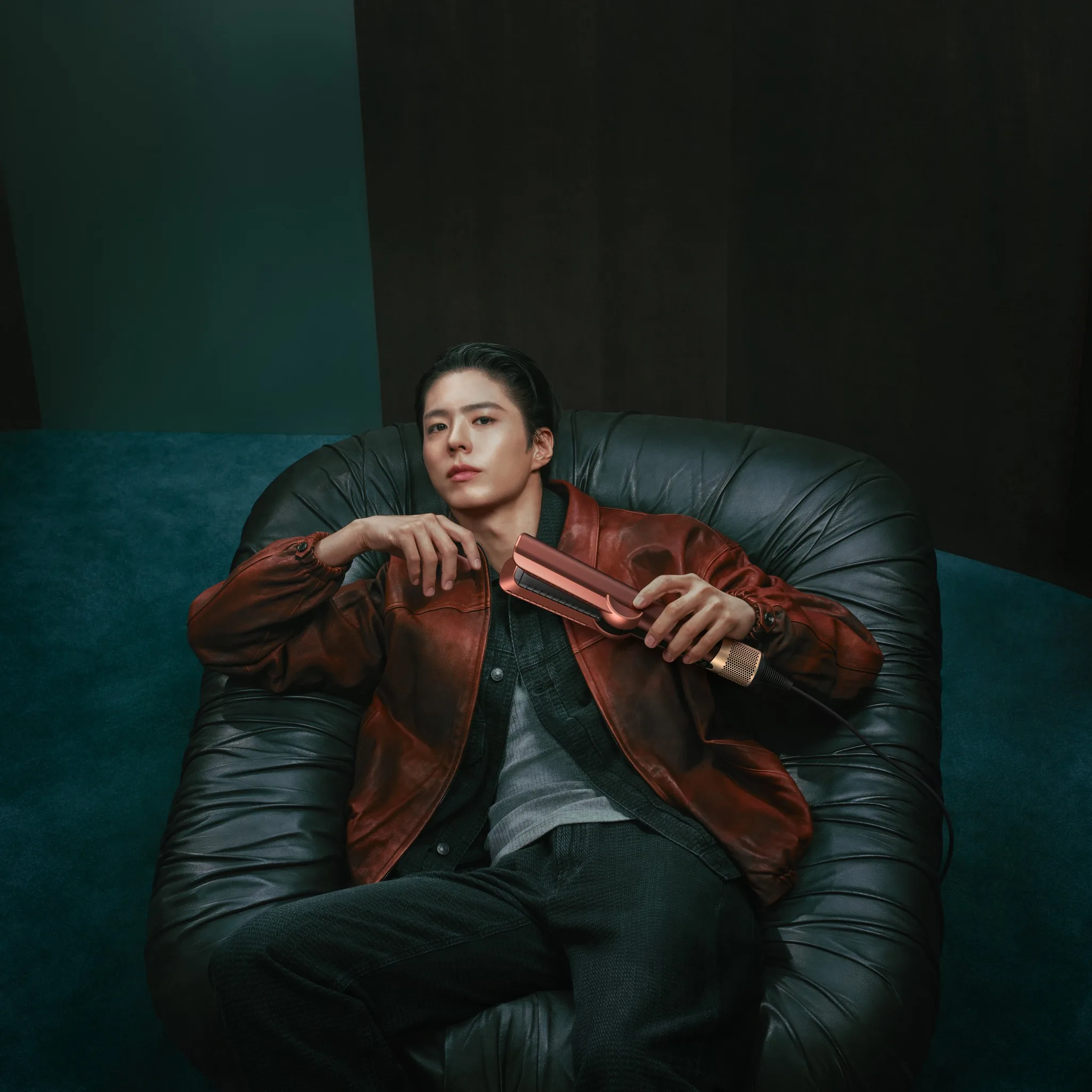 Park Bo Gum in a rust-red leather jacket holding a Plum and Copper Dyson device against a teal and dark background