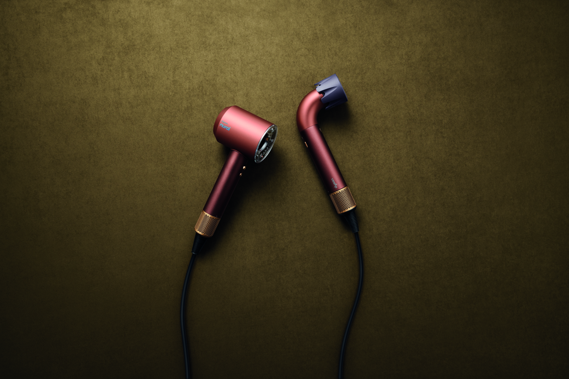 Two Dyson hair dryers in plum and copper tones with gold accents on a textured olive-green background