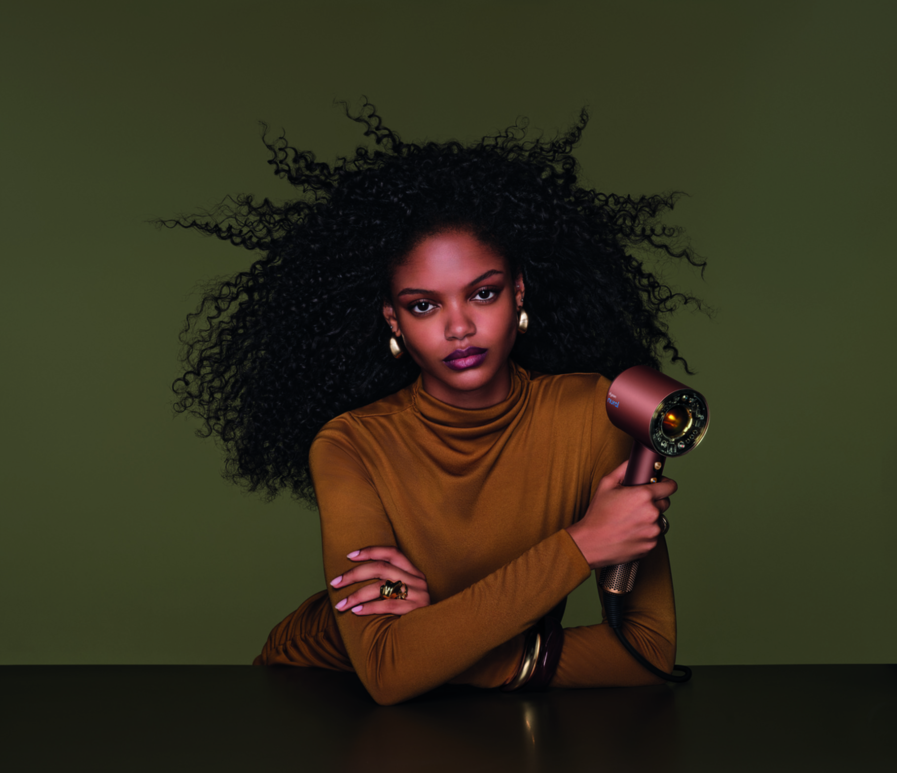 A person with long curly black hair in a bronze-toned shirt holds a Plum and Copper Supersonic hair dryer against a green background