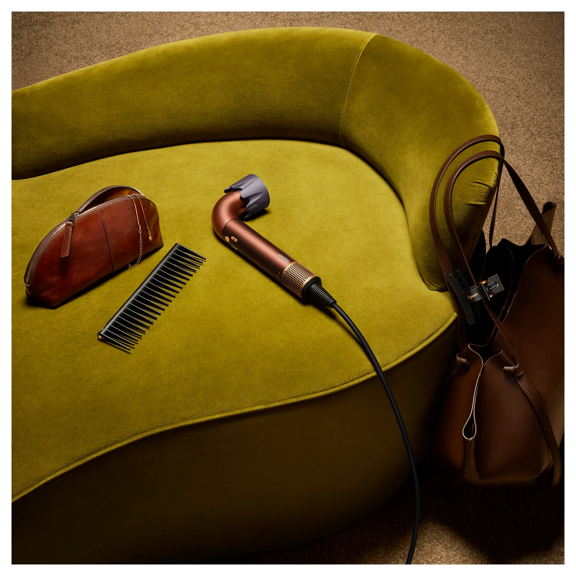 Dyson Supersonic r hair dryer in Plum and Copper on a chartreuse armchair with a comb, cosmetic pouch and crossbody bag