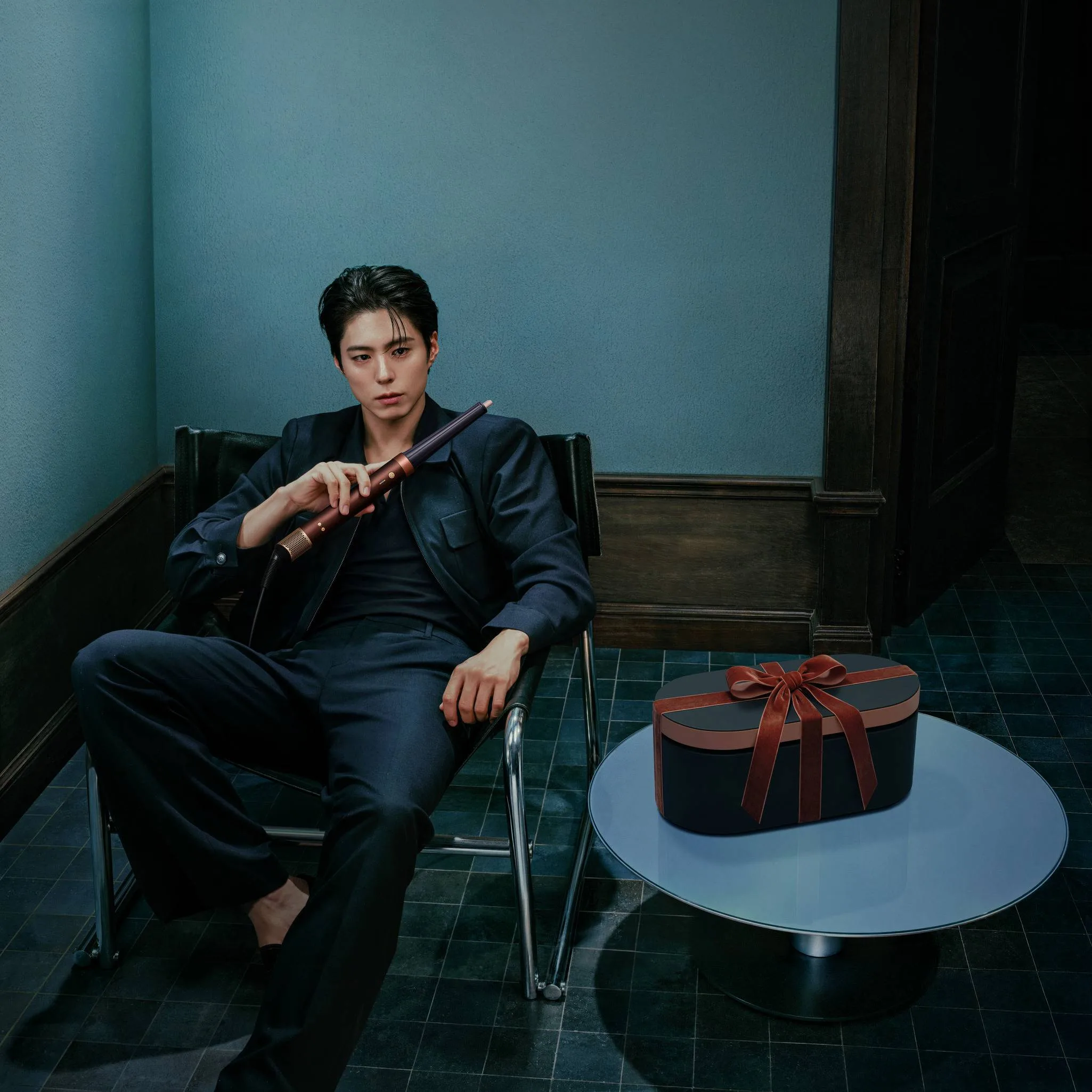 Park Bo Gum in a navy suit holding the conical barrel of a Plum and Copper Airwrap beside a heart-shaped gift box