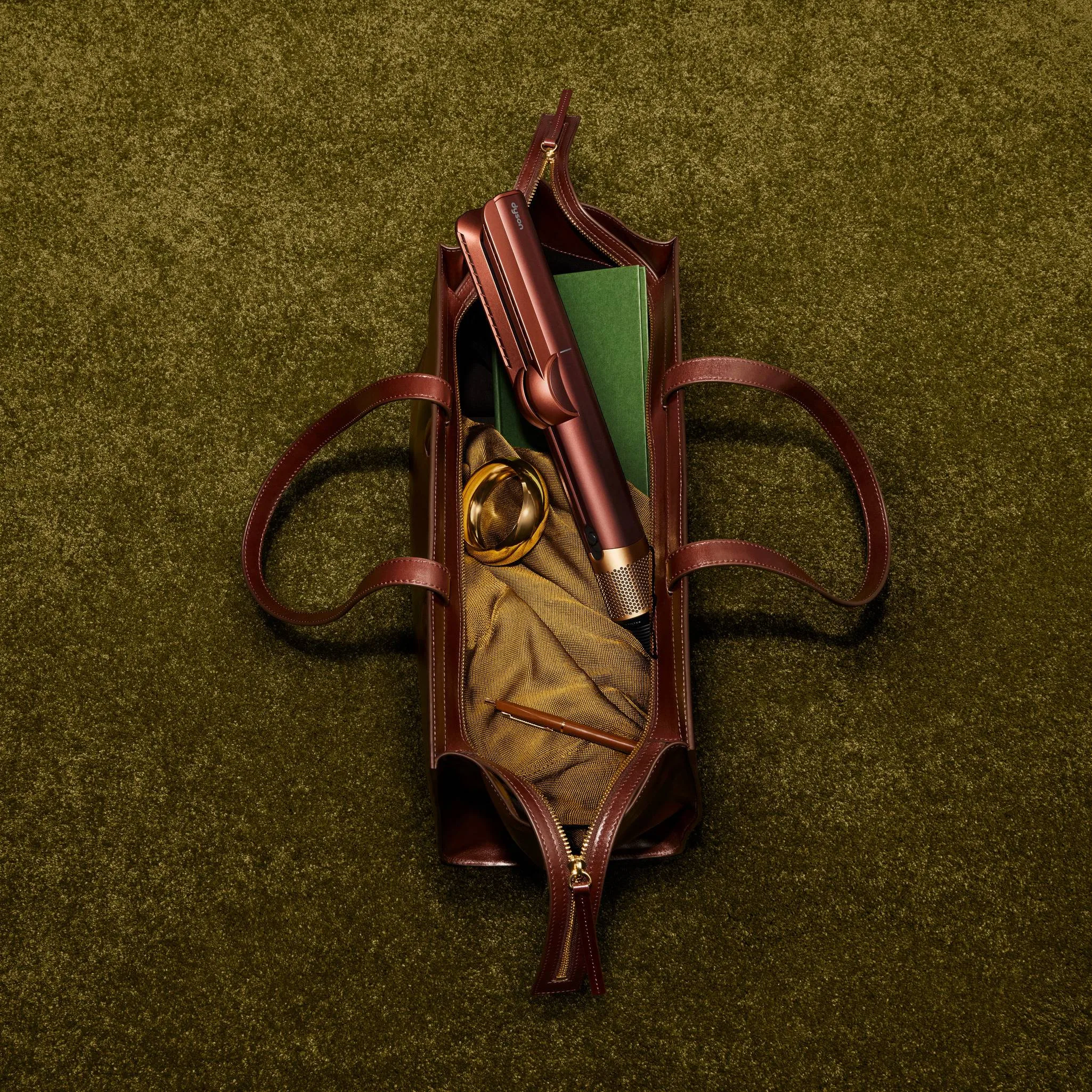An open Plum and Copper leather carry bag with a tan interior, gold-toned accessories and a green book on moss-green fabric