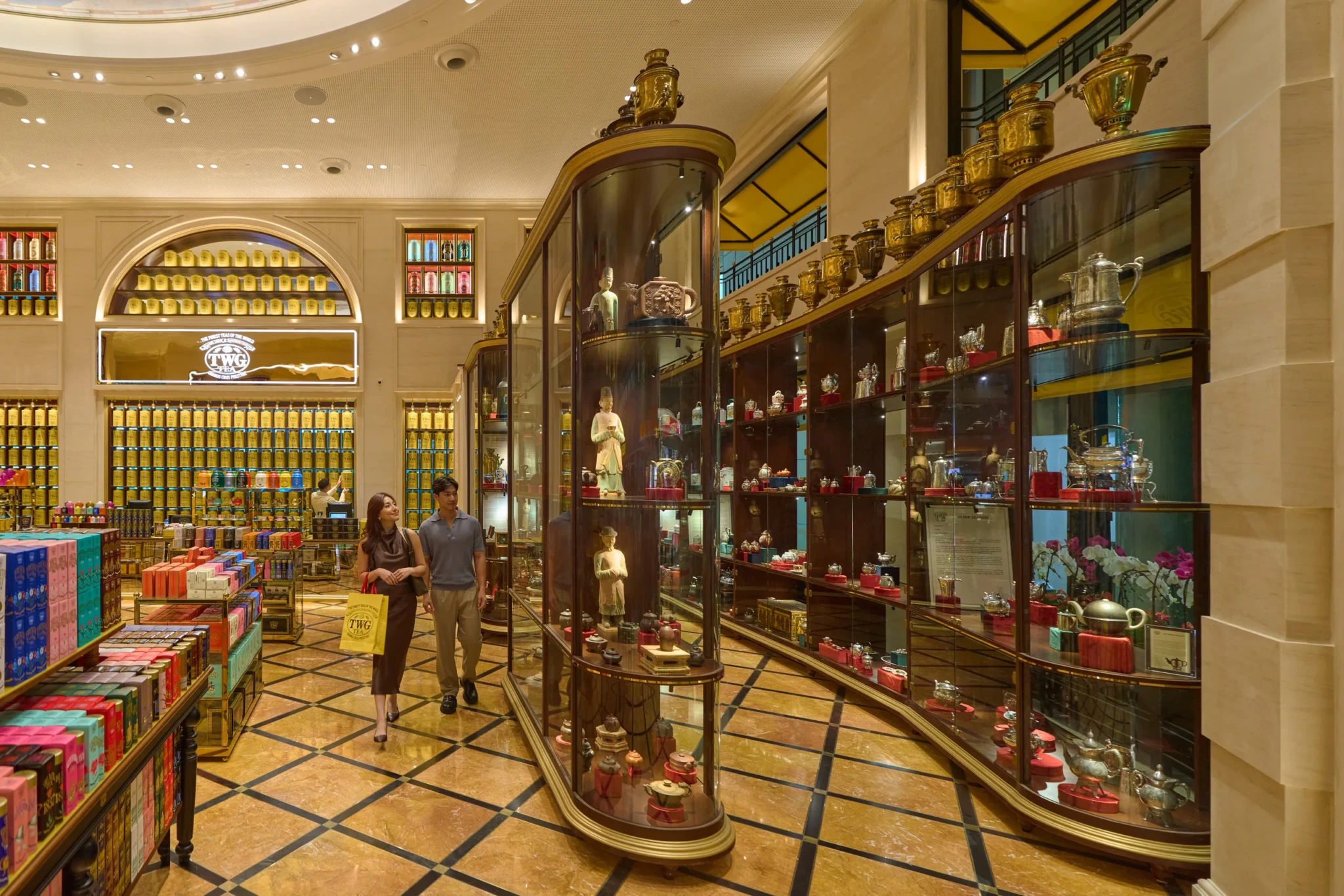 Interior of TWG Tea Museum with glass display cases featuring antique tea wares and Russian samovars, illuminated shelving with yellow tea canisters, and visito