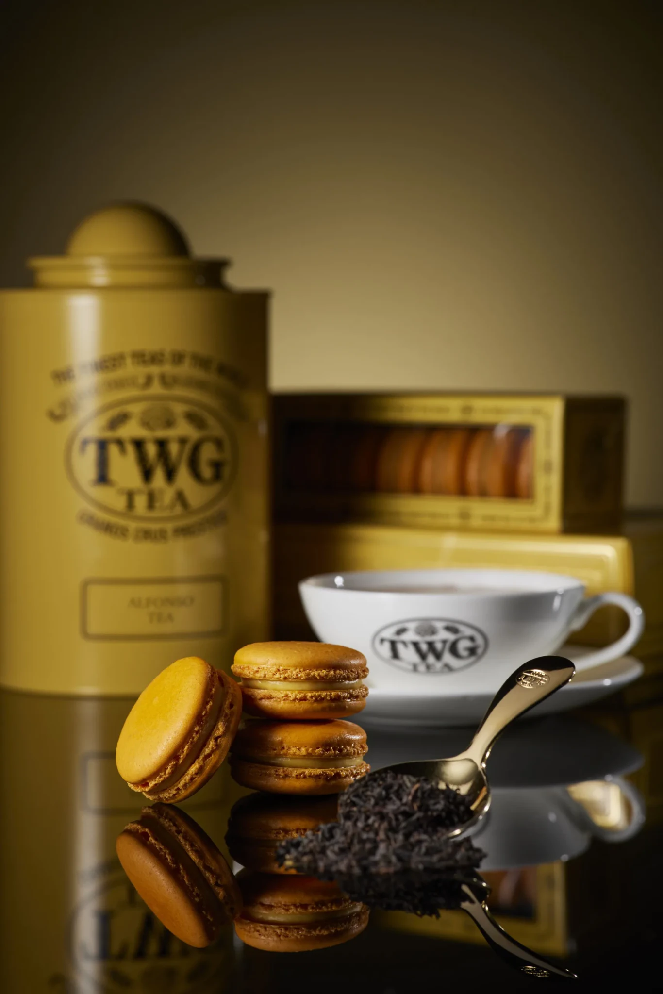Mango Macaron with Alfonso Tea-Infused Chocolate Ganache beside TWG Tea packaging, a porcelain teacup and loose tea leaves on a brass scoop