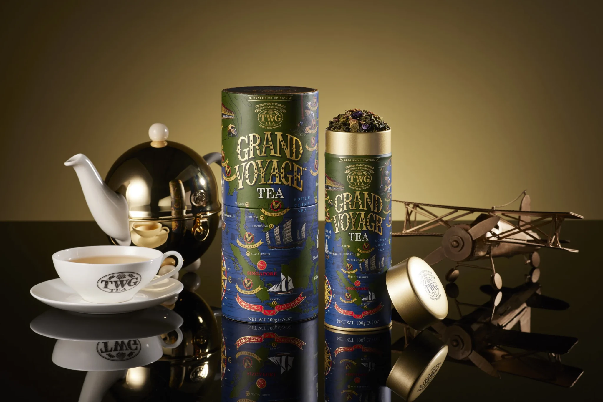 Grand Voyage Tea in its cylindrical Grand Mode Tea Collection packaging beside a porcelain teacup and teapot