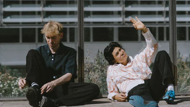 Aili & Orson sit together on a sunlit terrace with a modernist building behind them