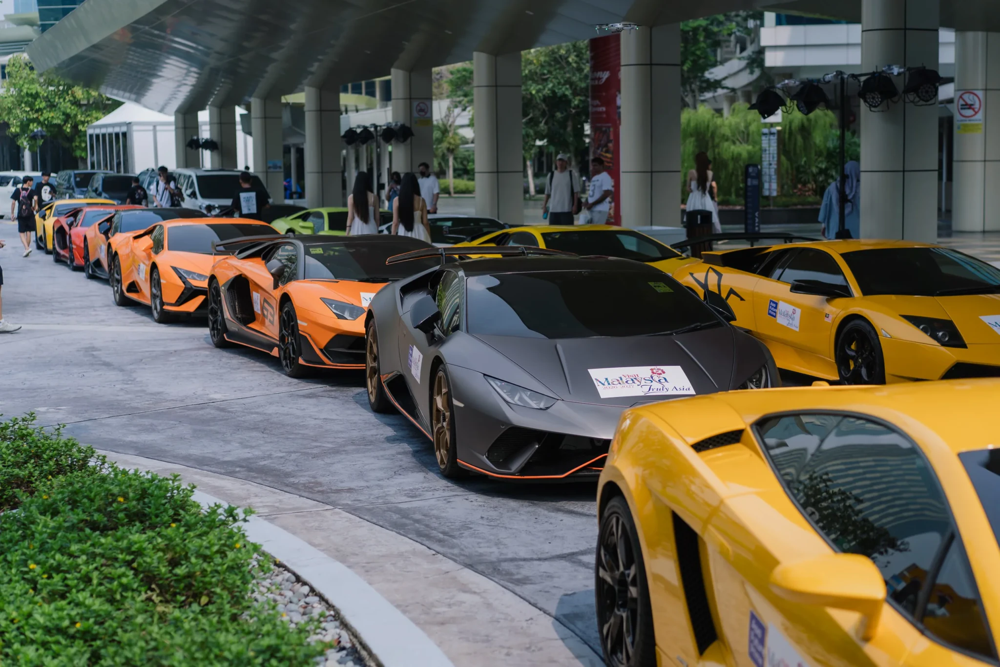 A row of orange and yellow Lamborghinis on display, with spectators and modern buildings behind them