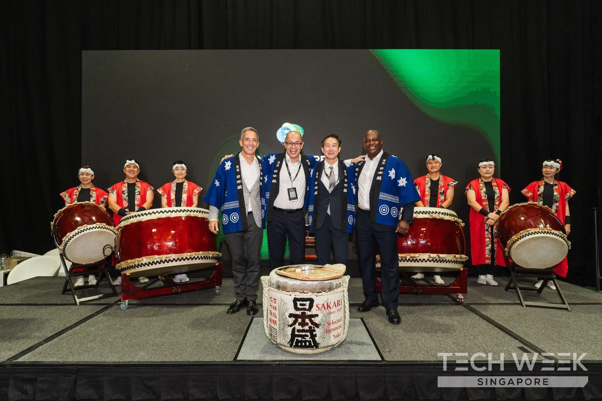 Four men in blue happi coats stand with taiko drummers behind a sake barrel on stage at Tech Week Singapore 2026