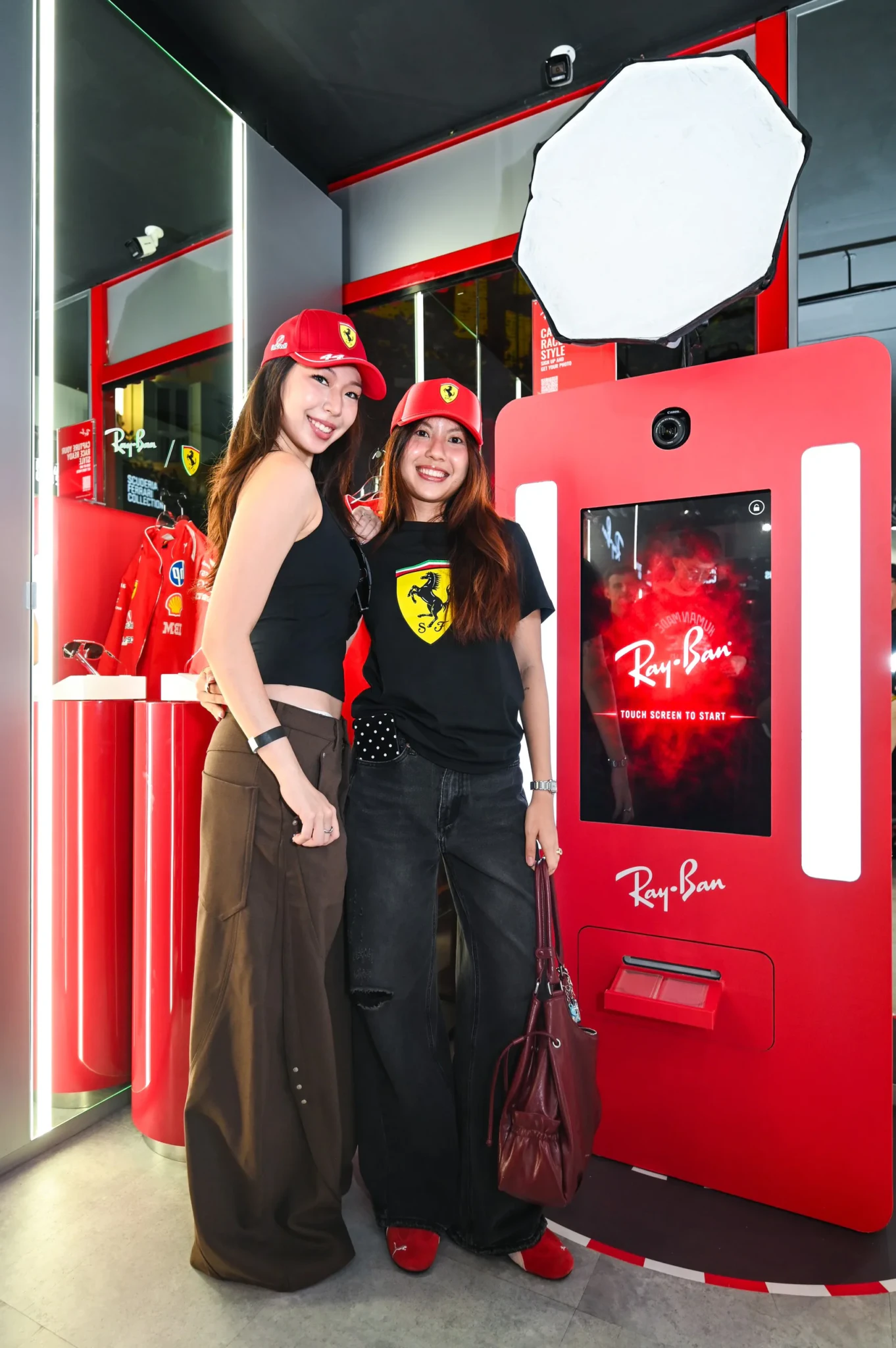 Two women in Ferrari caps posing among red display fixtures at the Ray-Ban pop-up