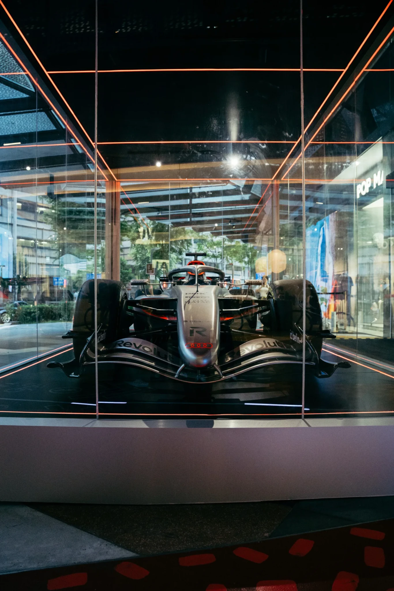 A Formula 1 show car in black and silver livery behind glass with orange neon framing at the Off The Grid pop-up at Funan