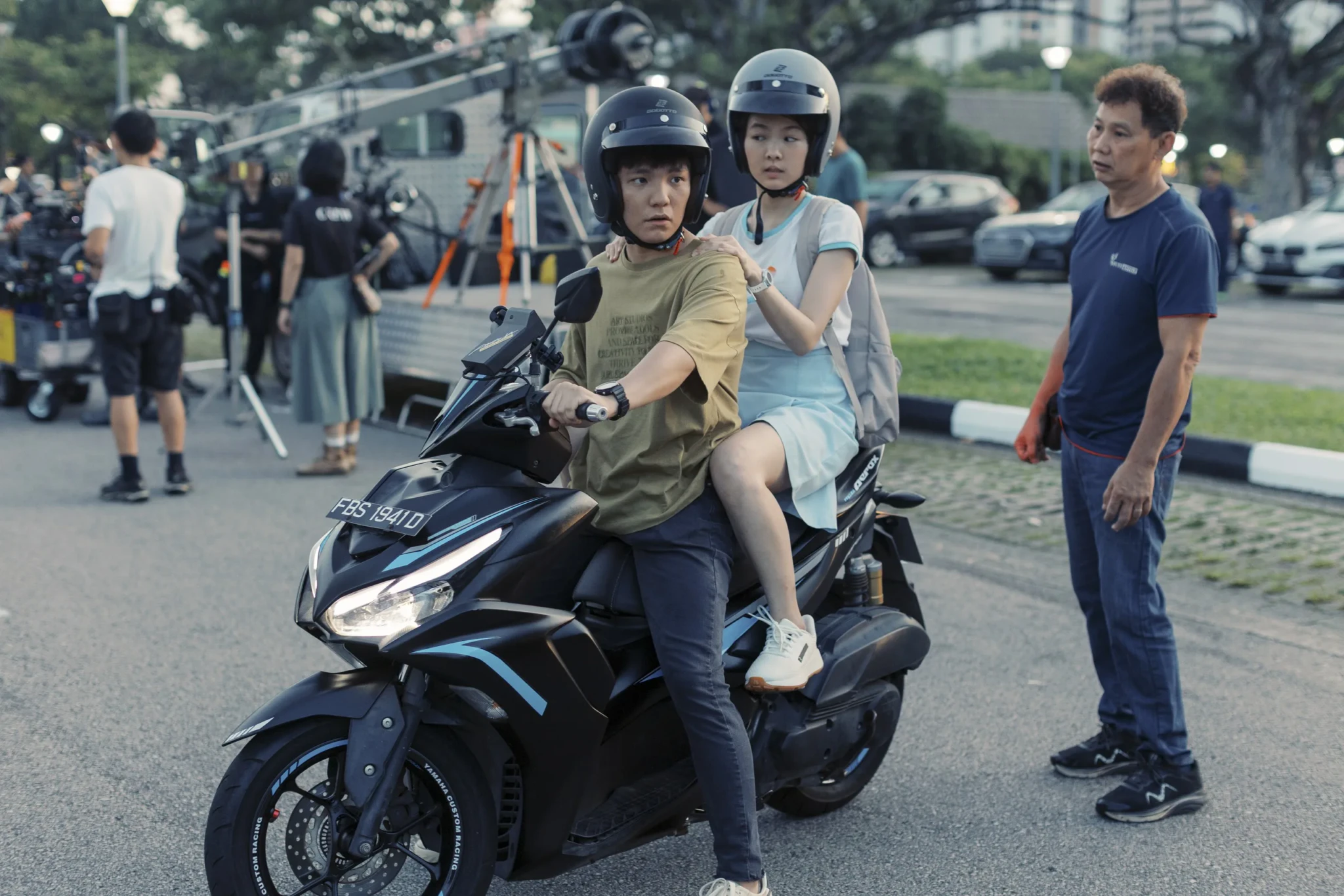Two people in helmets on a scooter with production crew in the background