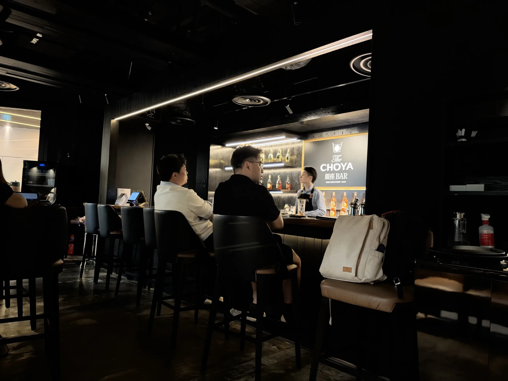 Bar interior with guests seated at counter and bartender, illuminated CHOYA bottle display behind