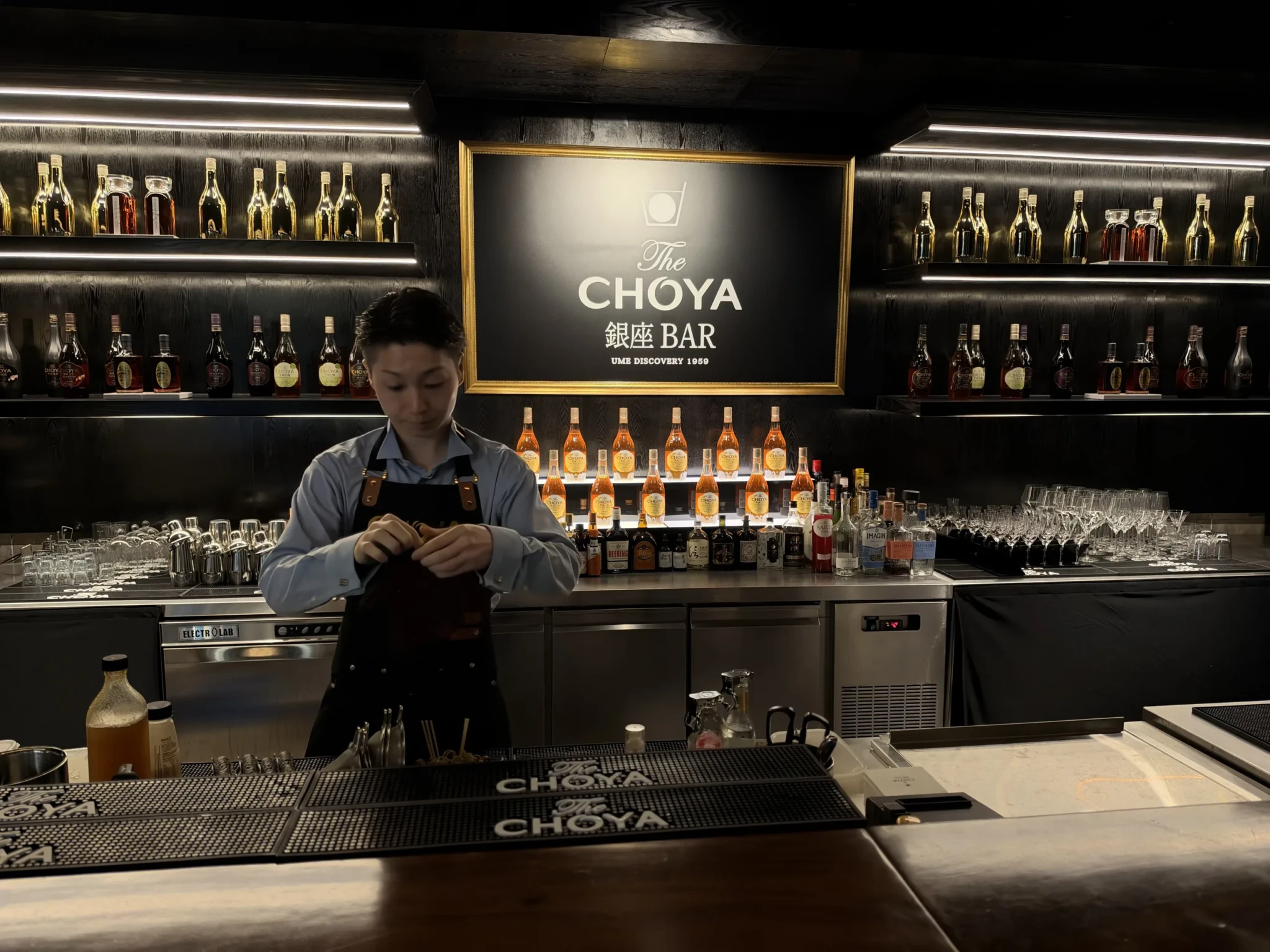A bartender working at the counter with illuminated shelves of Japanese spirits and glassware behind
