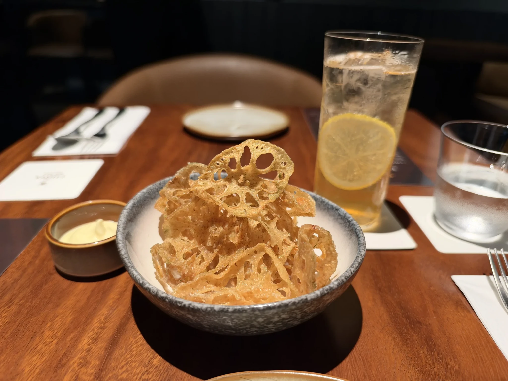 Crispy lotus root chips piled in a textured grey bowl beside a glass of lemon water and place settings