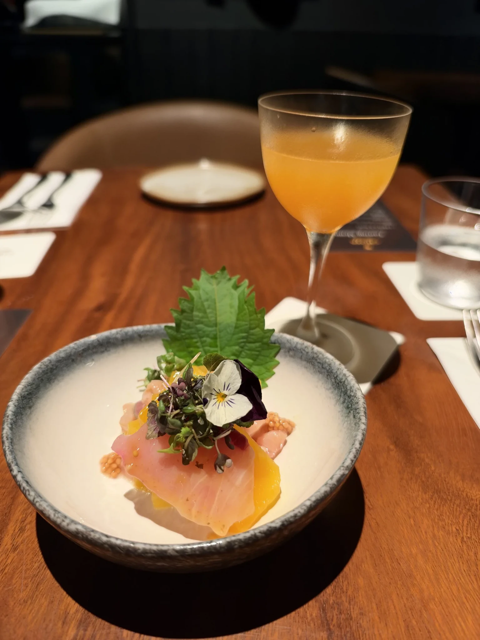 Sliced fish with edible flowers and microgreens in a ceramic bowl, served next to a CHOYA ume cocktail