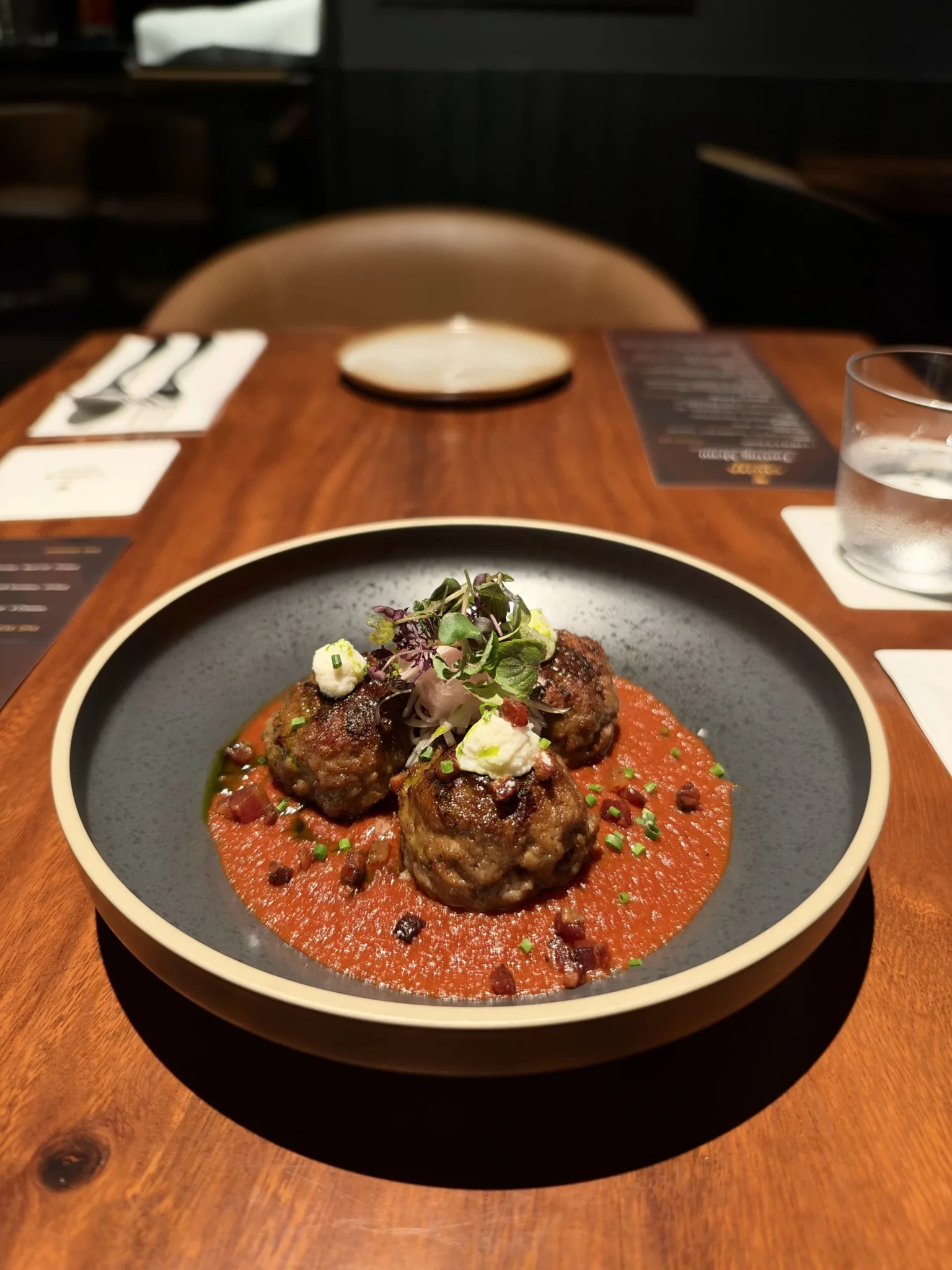 Three meatballs in red sauce topped with microgreens and white cheese foam on a grey plate