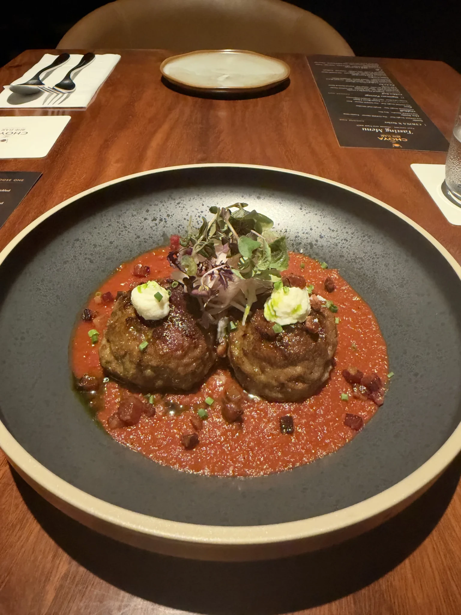 Two brown meatballs on a grey plate with red tomato sauce, garnished with microgreens and white cream