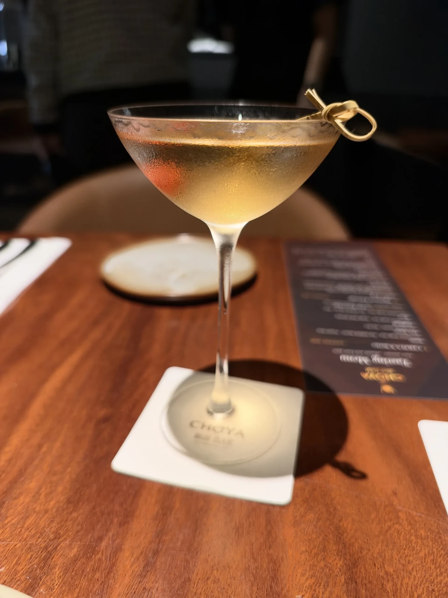 A CHOYA ume cocktail in a coupe glass with a gold ring charm on a white square coaster