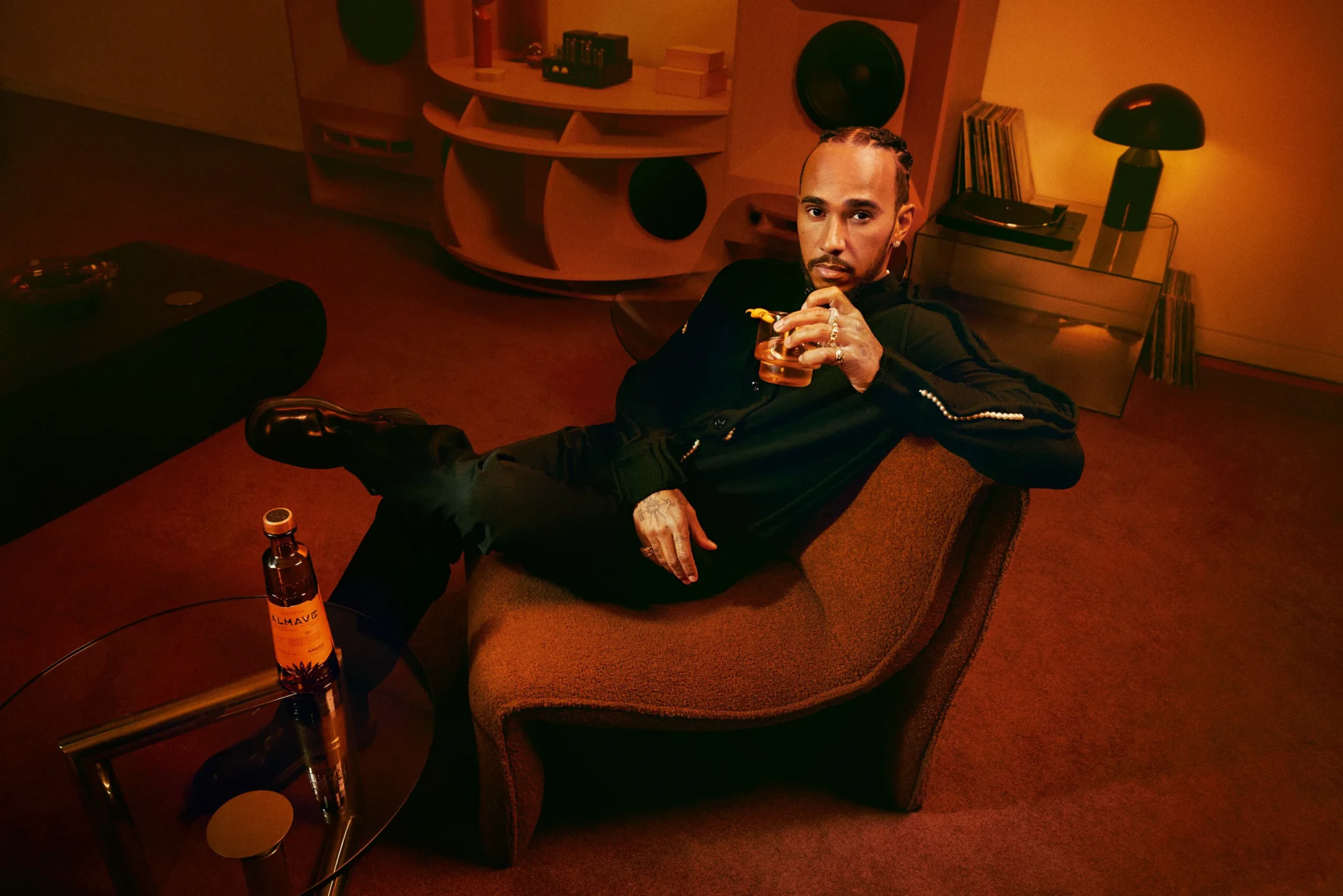 Lewis Hamilton seated in a modern chair holding a glass of Almave spirits in an orange-lit interior