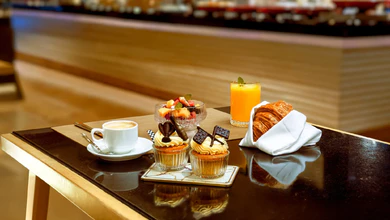 A breakfast spread at Oscar's with a cappuccino, fruit salad, two decorated cupcakes, a croissant in white linen, and orange juice on a dark table
